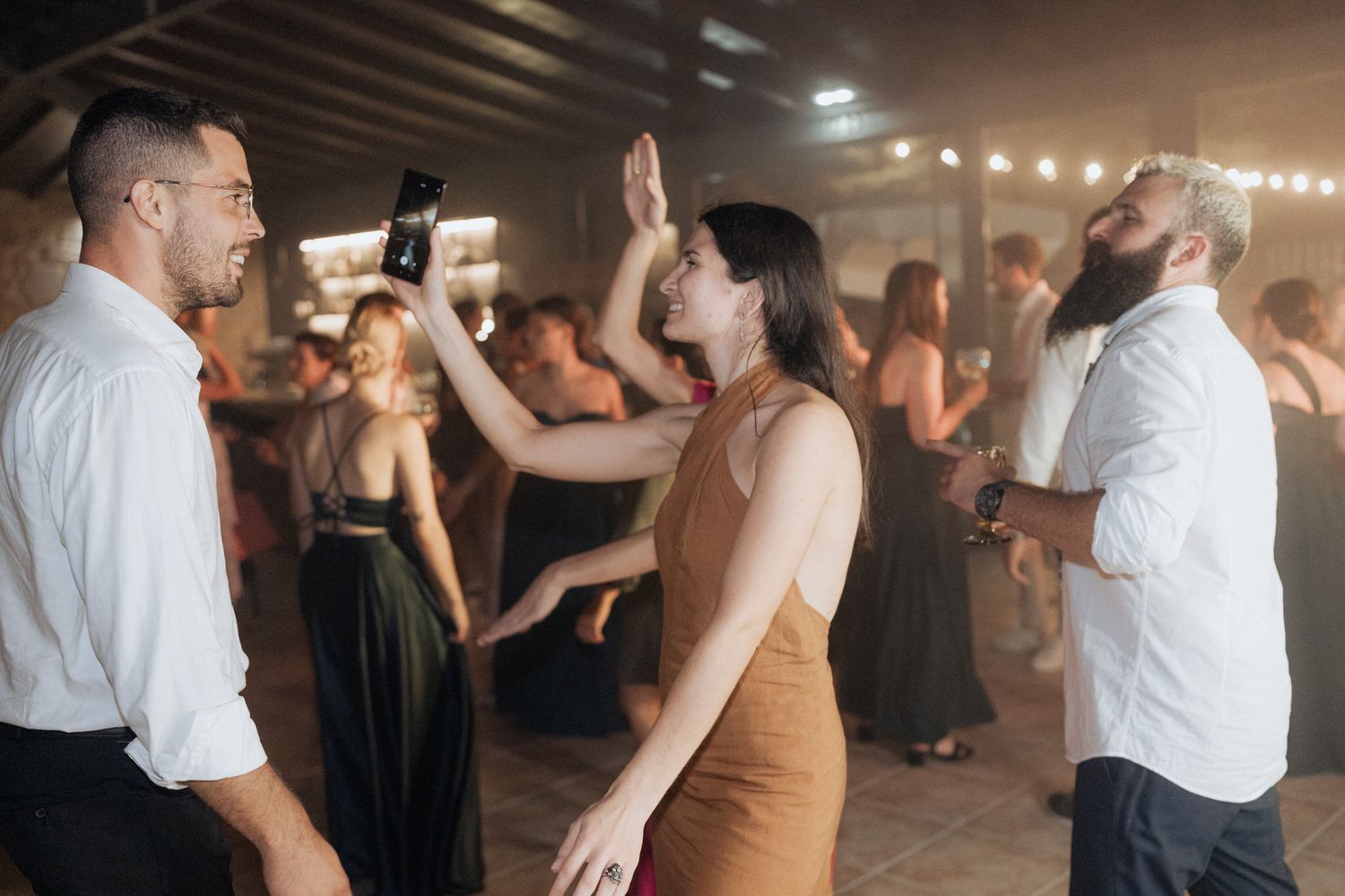 Dancing guests celebrate at a lively indoor reception with decorative lighting overhead.