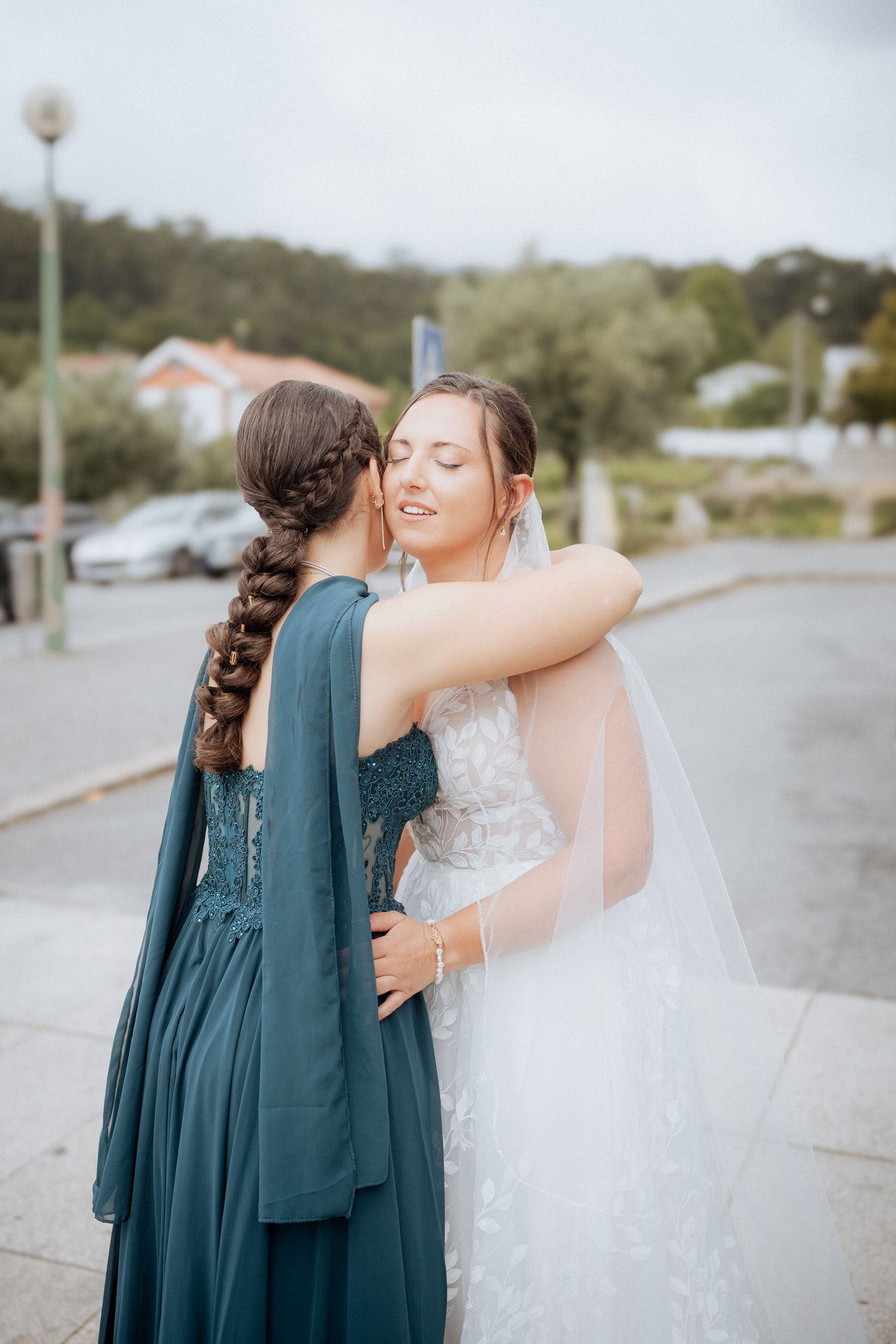 A tender moment between a bride in white and a bridesmaid in a teal dress sharing an embrace outdoors.