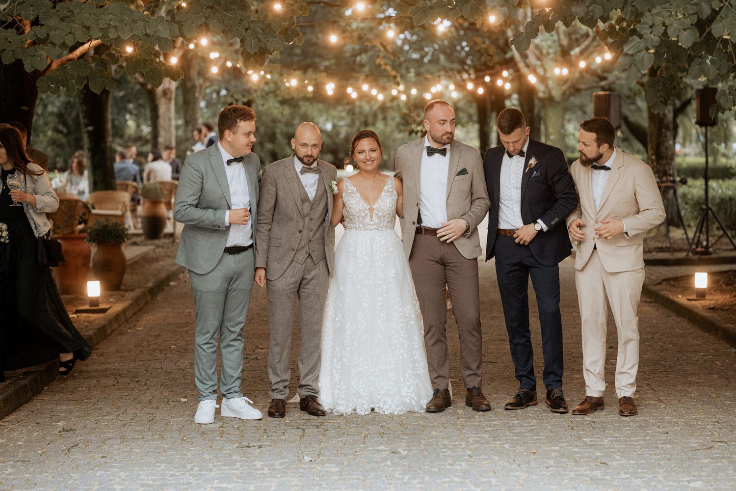 A wedding party stands together outdoors at night under glowing string lights with candle-lined path.