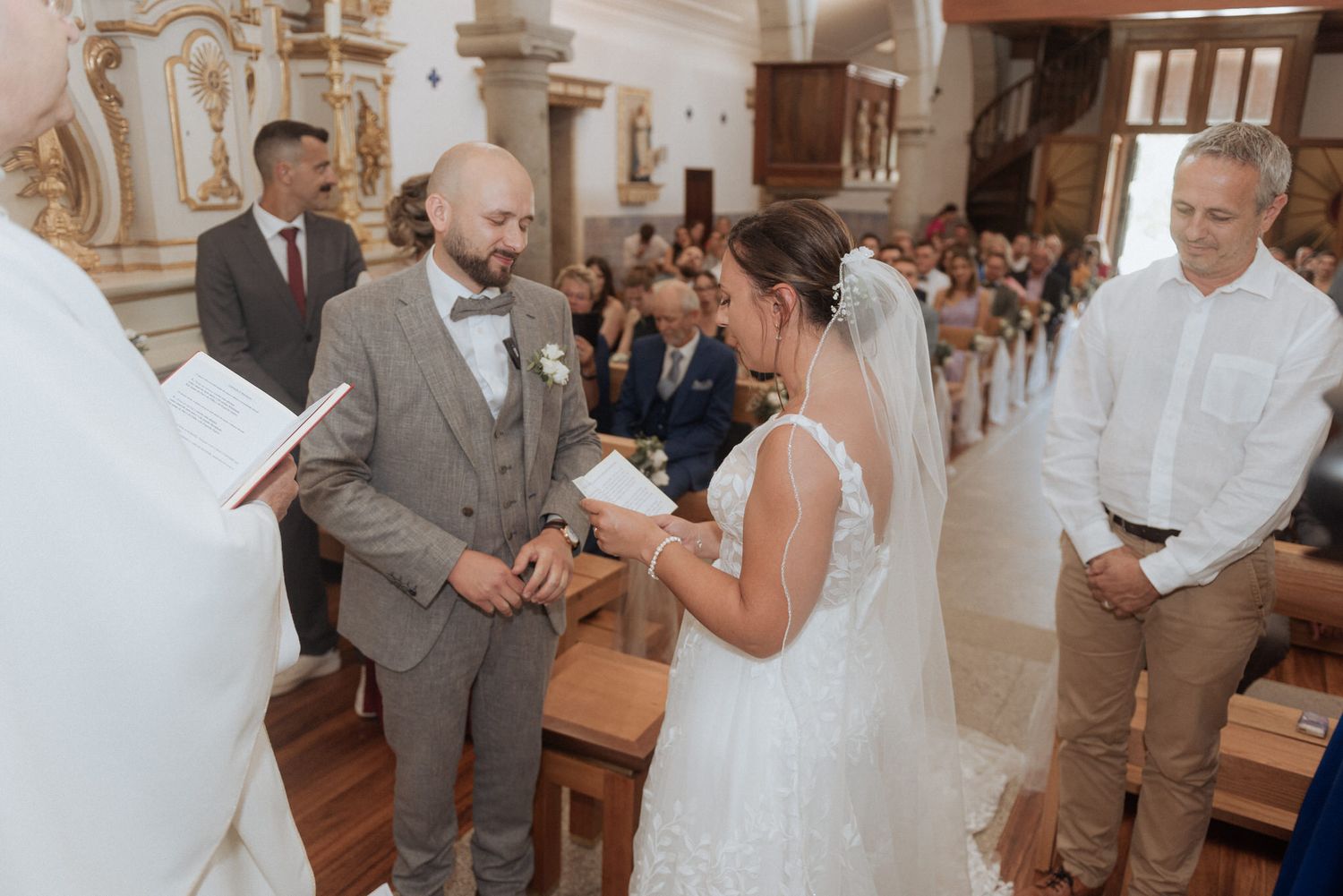 A couple exchanges vows during their wedding ceremony in an ornate church interior surrounded by loved ones.