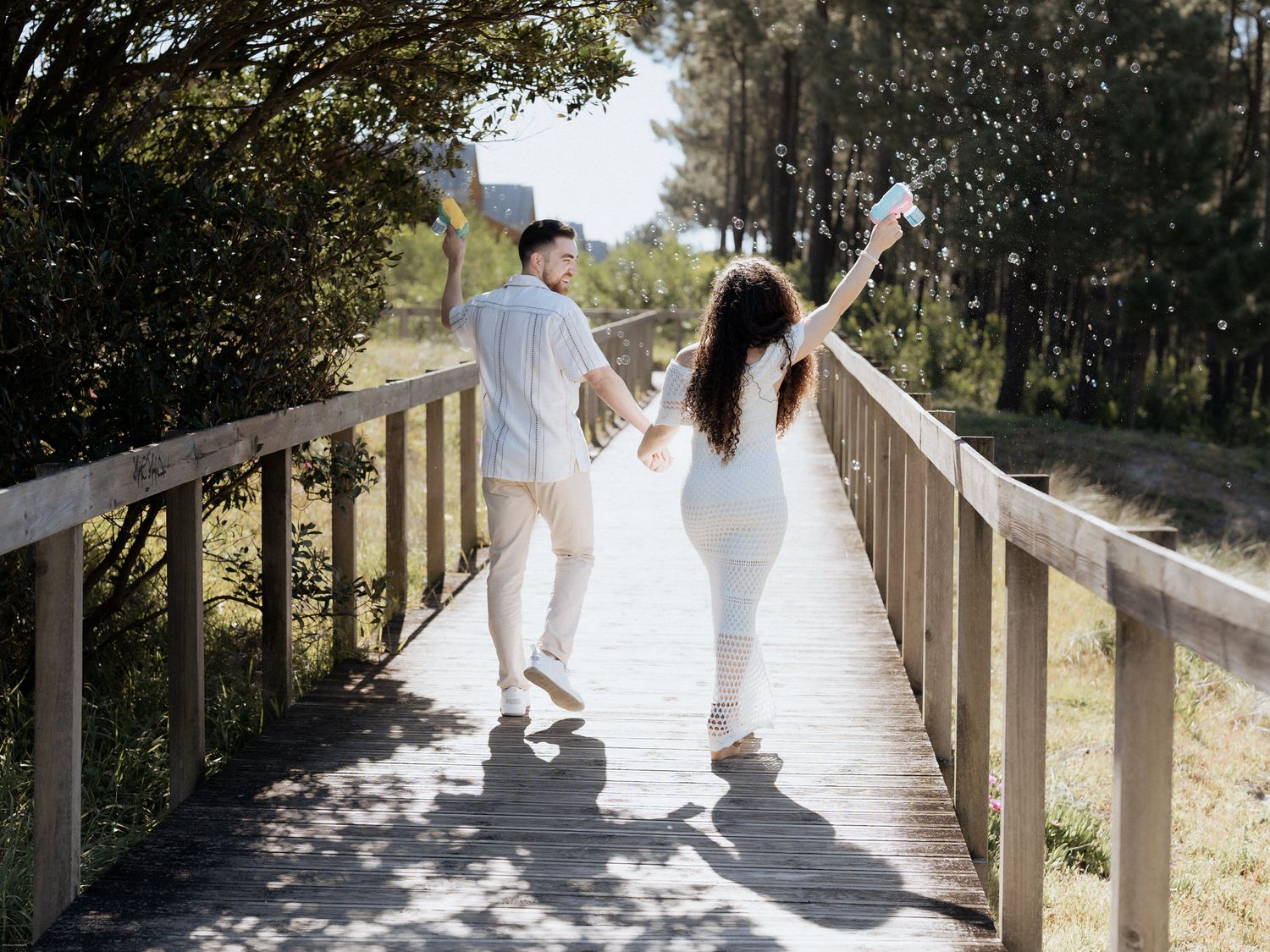 Um casal em trajes brancos combinando comemora alegremente com os braços erguidos em um calçadão de madeira cercado pela natureza.