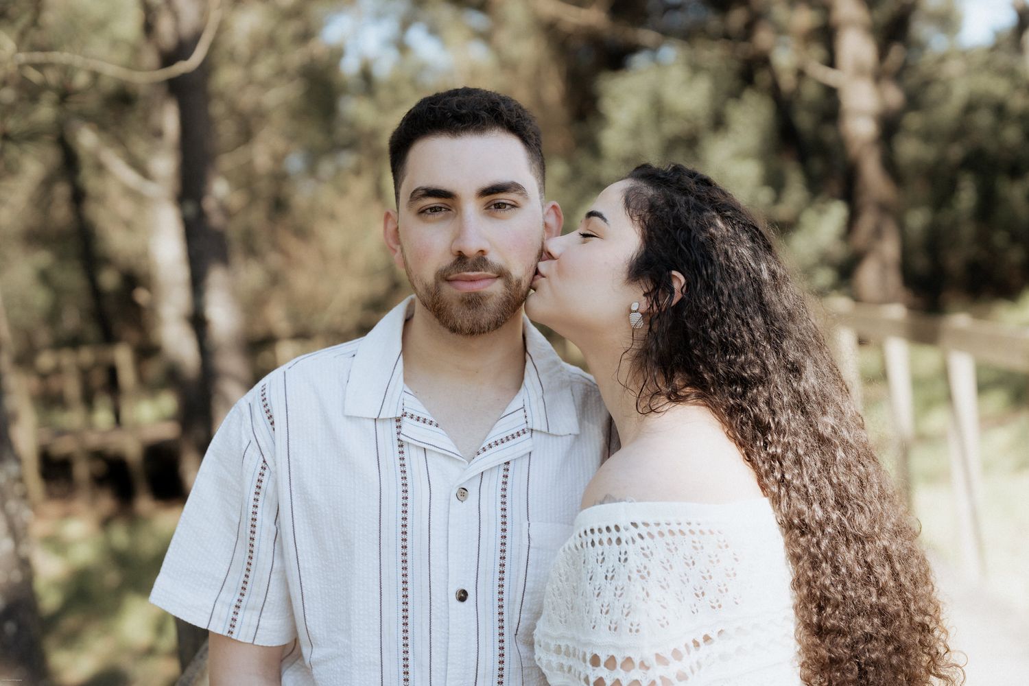 Um casal partilha um momento de ternura ao ar livre, com a luz natural do sol filtrada pelas árvores ao fundo.