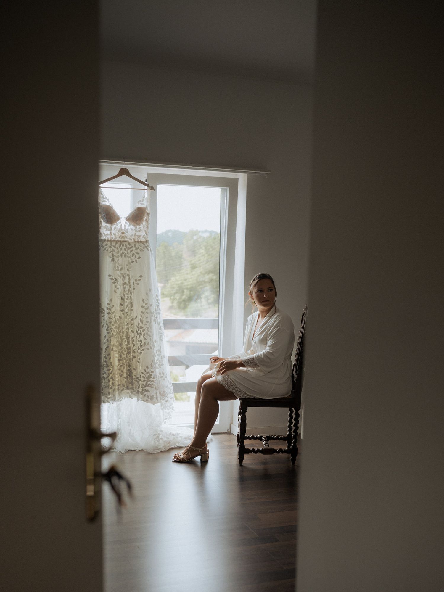 A wedding dress hangs by a window as someone in a white robe sits quietly in a chair, creating a serene bridal preparation scene.