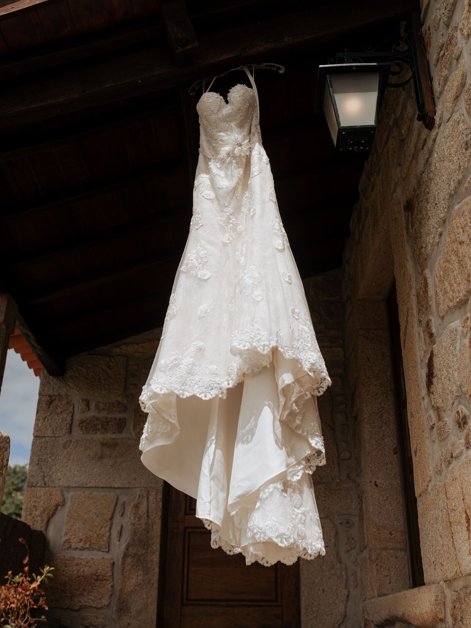 A series of white lace wedding dresses hang in an archway of a rustic stone building in natural light.