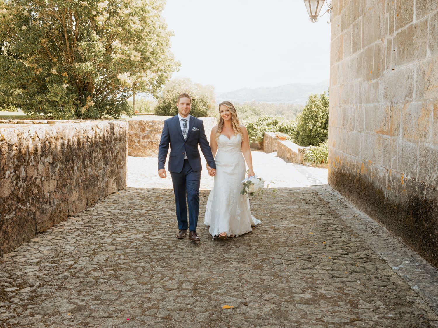 A couple walks down a scenic cobblestone path between stone walls on their wedding day.