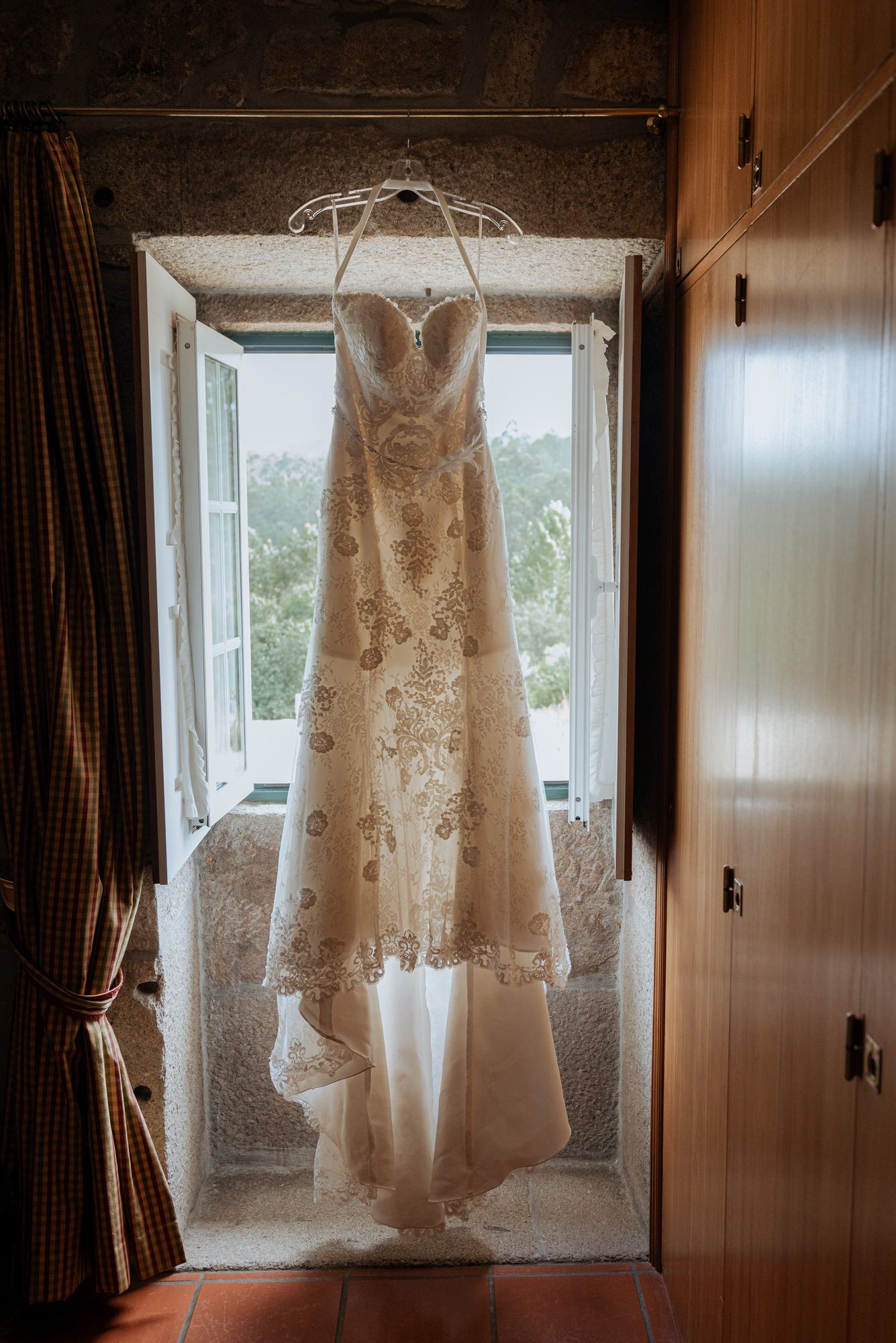 Wedding dress hangs gracefully against backlit window in warm light creating ethereal silhouette display.