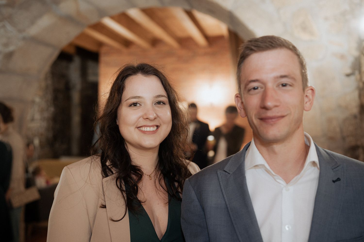 Young couple dressed in business casual attire smile warmly in an indoor venue with stone archways and warm lighting.
