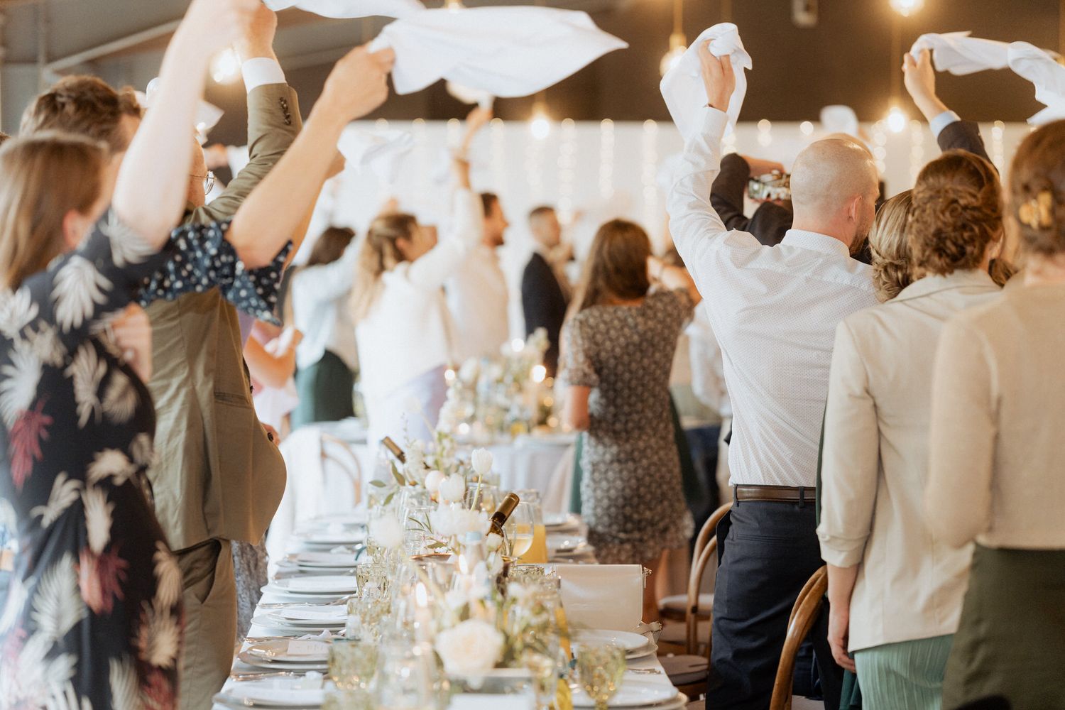 Wedding guests wave white napkins at a reception dinner with elegant table settings and floral centerpieces.