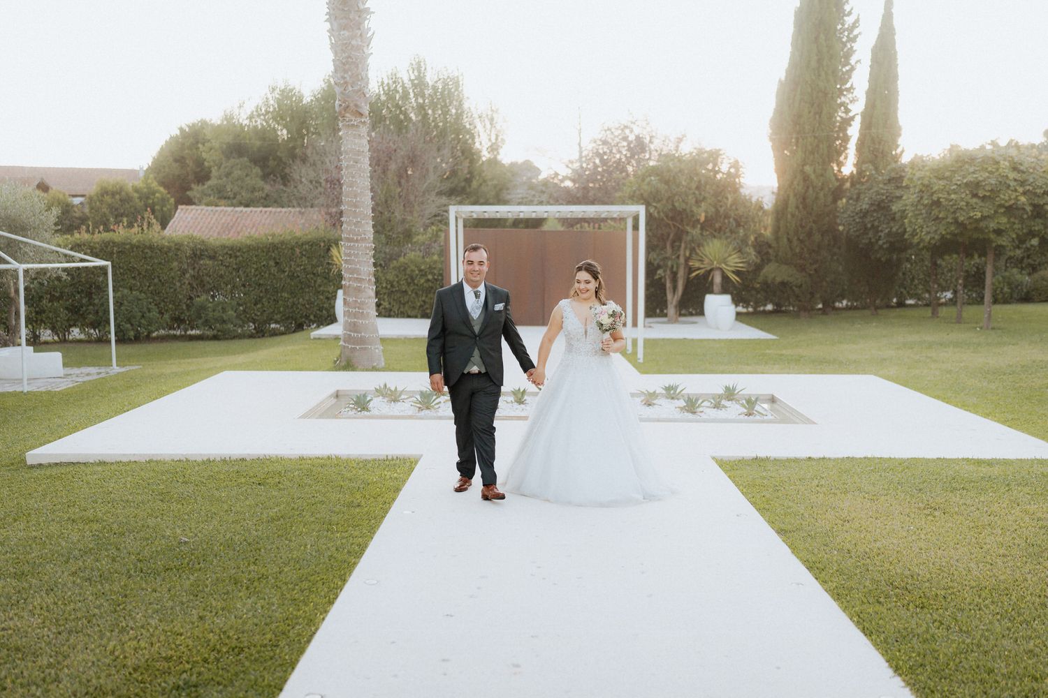 Two wedding figures walk down an outdoor white aisle runway with a modern backdrop and lush green lawn surroundings.