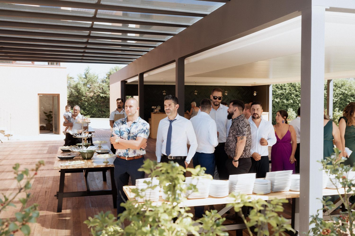 Wedding guests mingle and socialize beneath an outdoor covered patio with wooden beams during a daytime reception.