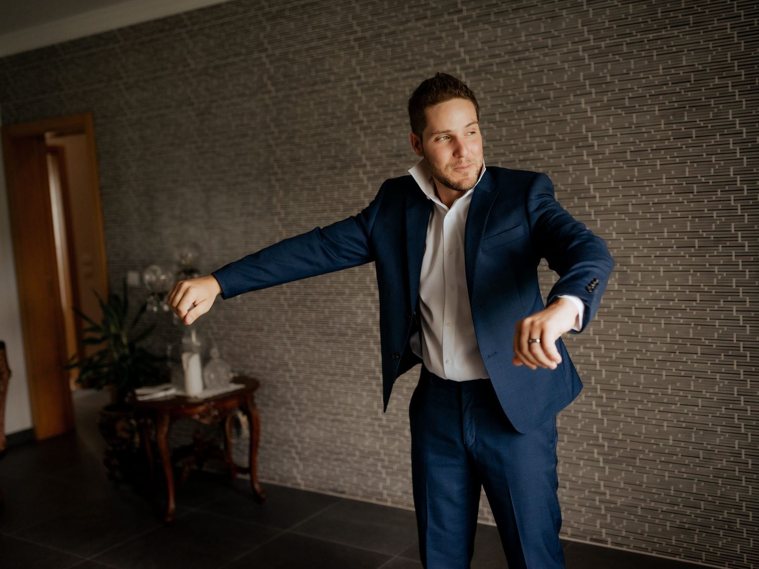 A person in a navy blue suit strikes a playful pose against a textured wall in an interior setting.