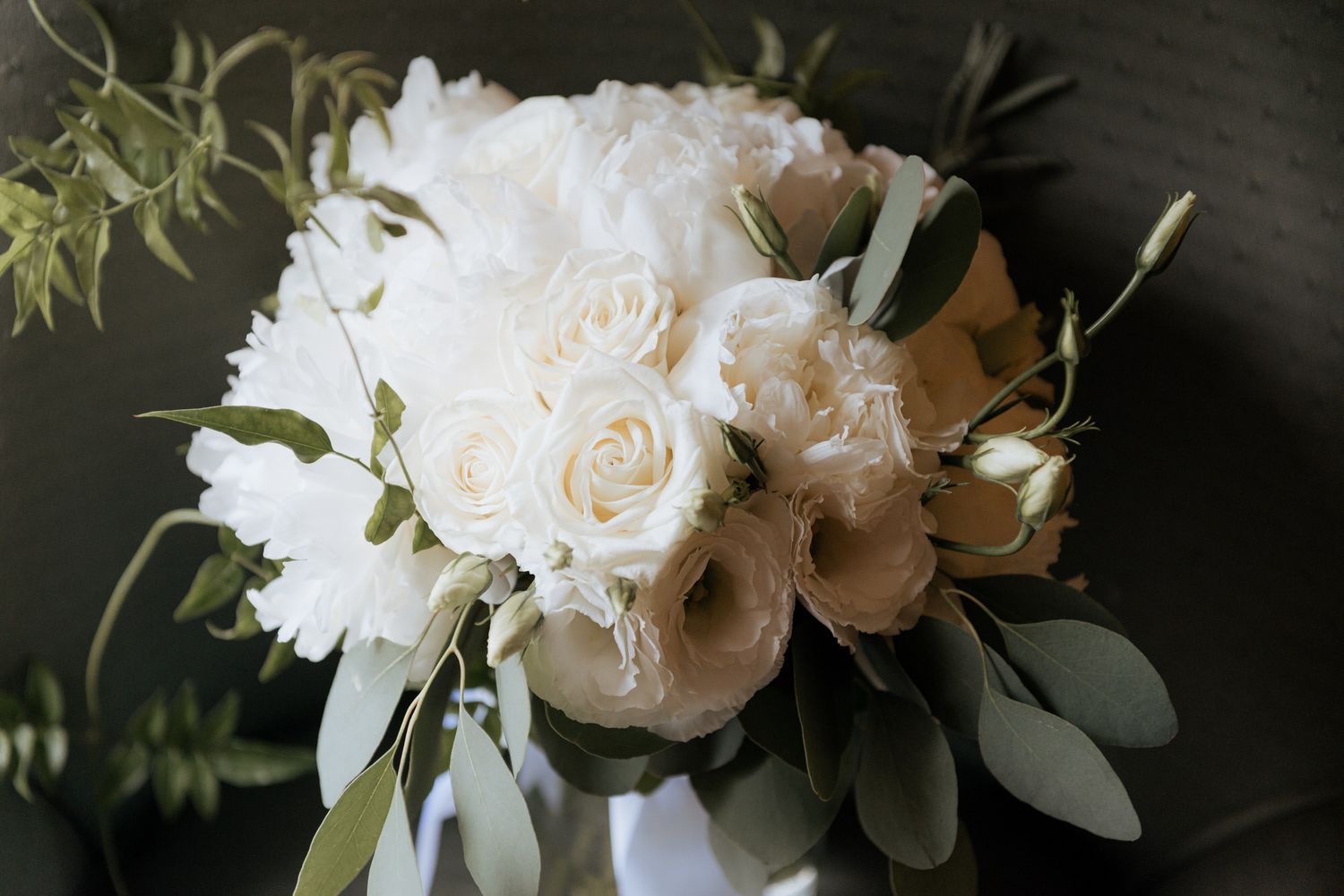 A romantic white wedding bouquet with roses, peonies and eucalyptus leaves against a dark background.