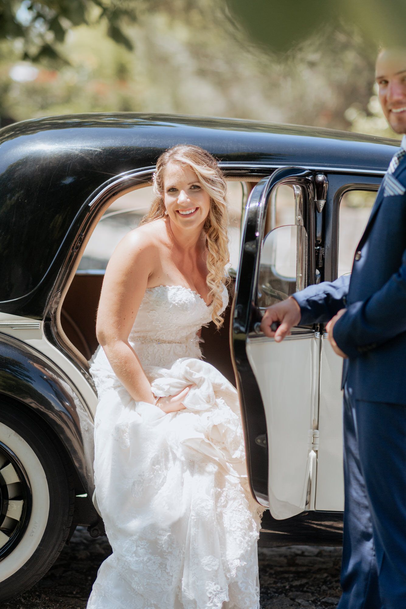 A bride in a white wedding dress gets into a vintage car at her wedding ceremony.