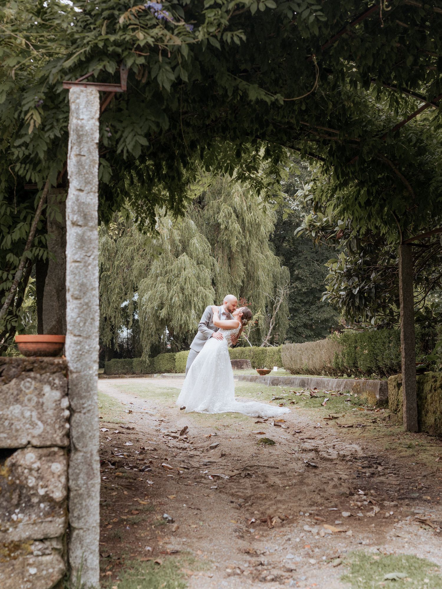 A romantic garden photo showing white wedding attire amid stone pillars and weeping willow trees on a dirt path.