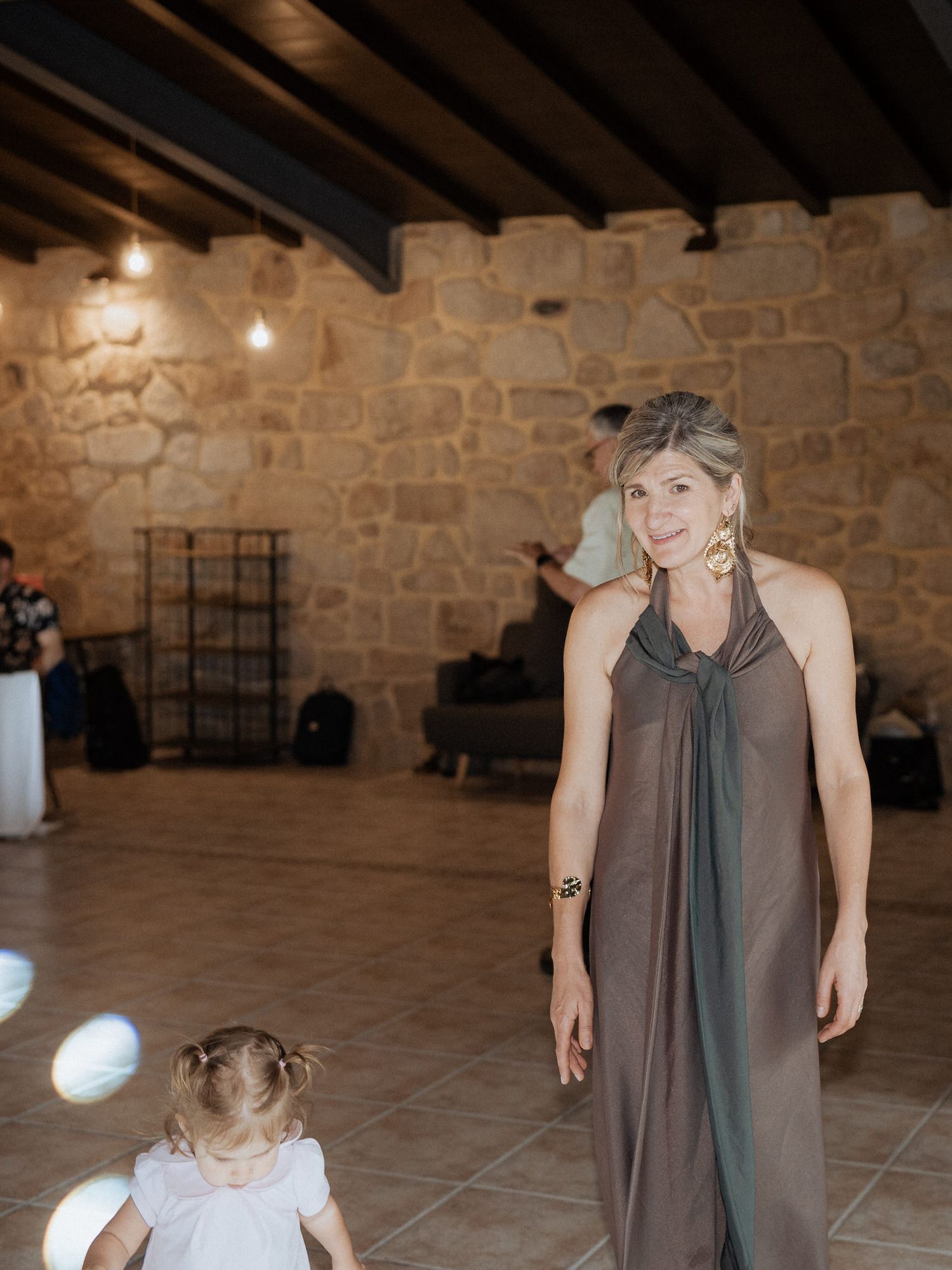 A person in a flowing gray halter dress stands in a rustic stone-walled room with wooden beams while a child looks on.