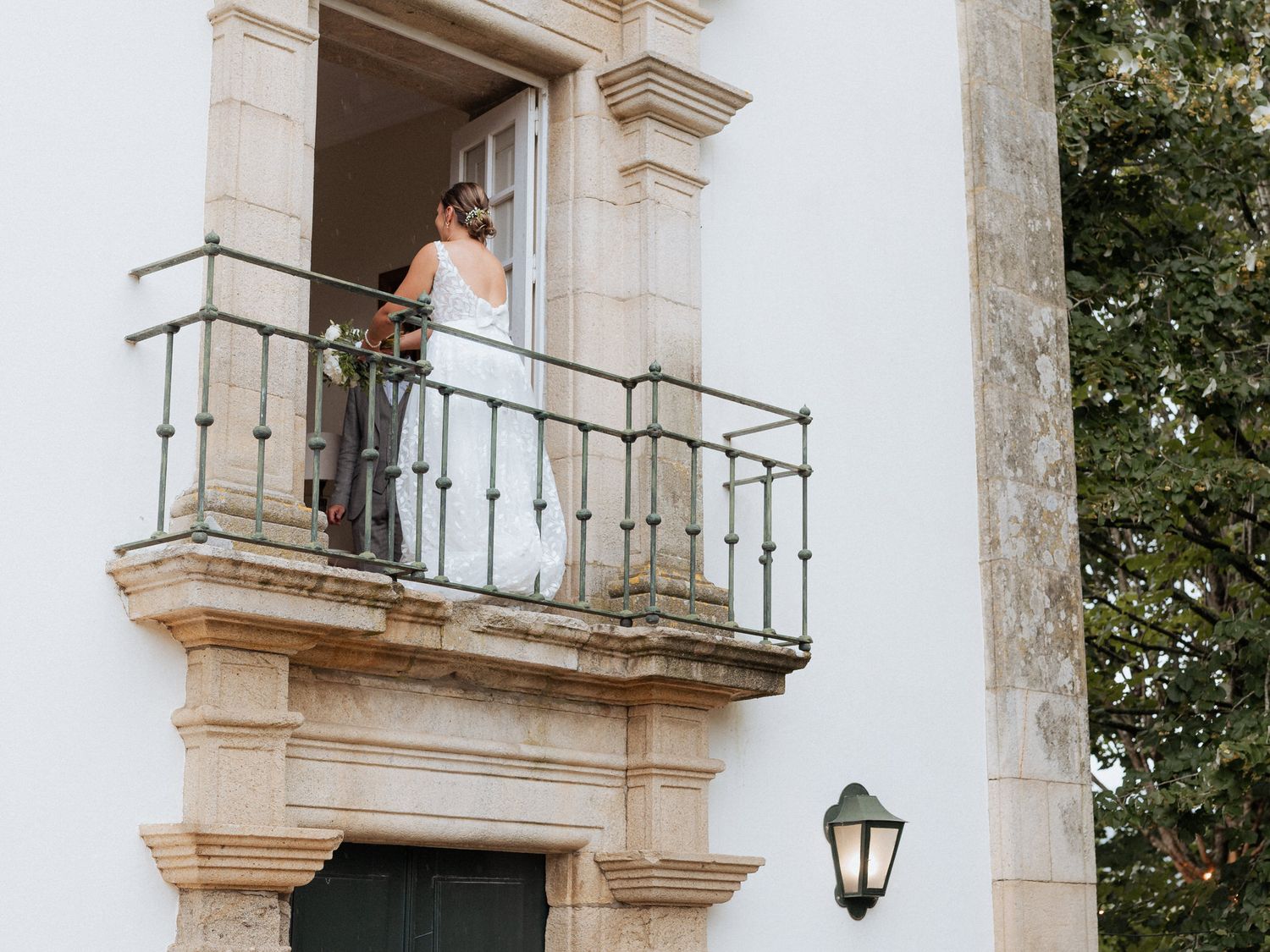 A couple stands on an elegant stone balcony with ornate railings overlooking a garden.
