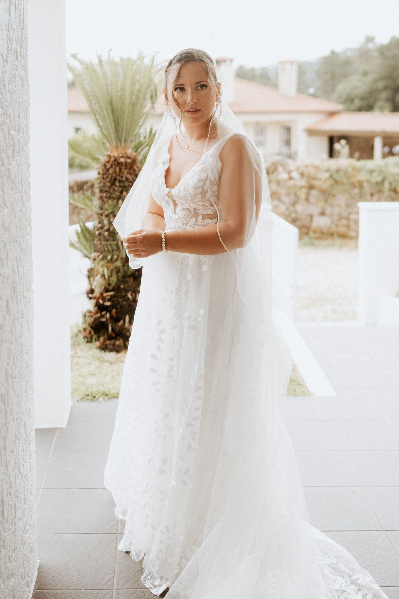 A bride in a flowing lace wedding gown with long veil poses elegantly on a white porch with tropical plants in background.