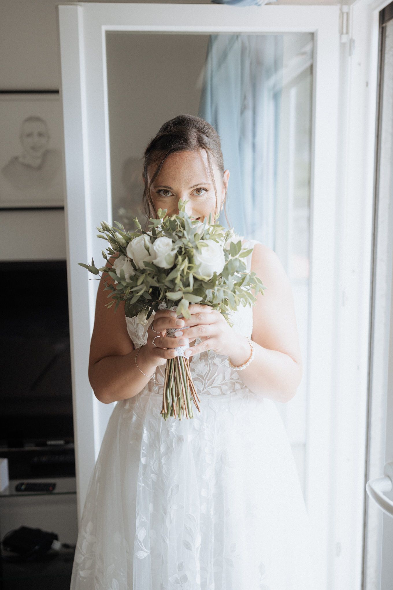 A bride in a white lace dress holds a delicate bouquet of white roses and eucalyptus by a window.