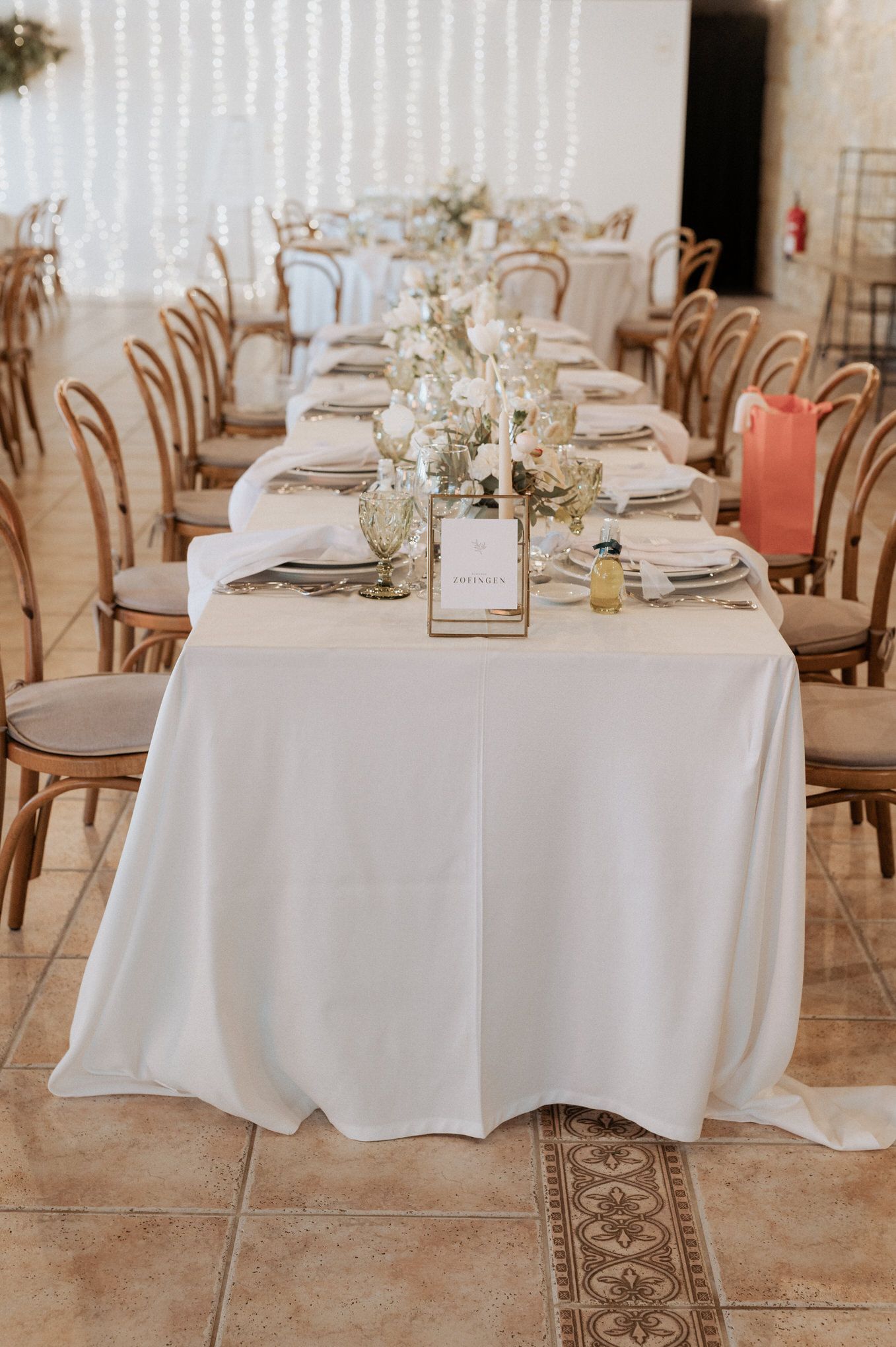 An elegant wedding reception table setting with wooden chairs and white linens in a naturally lit venue.