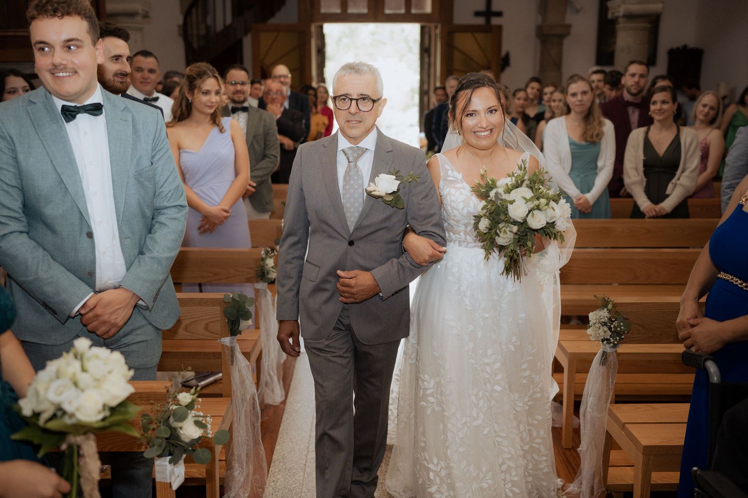 A bride in a sparkling gown walks down the church aisle accompanied by an elderly gentleman in a grey suit.