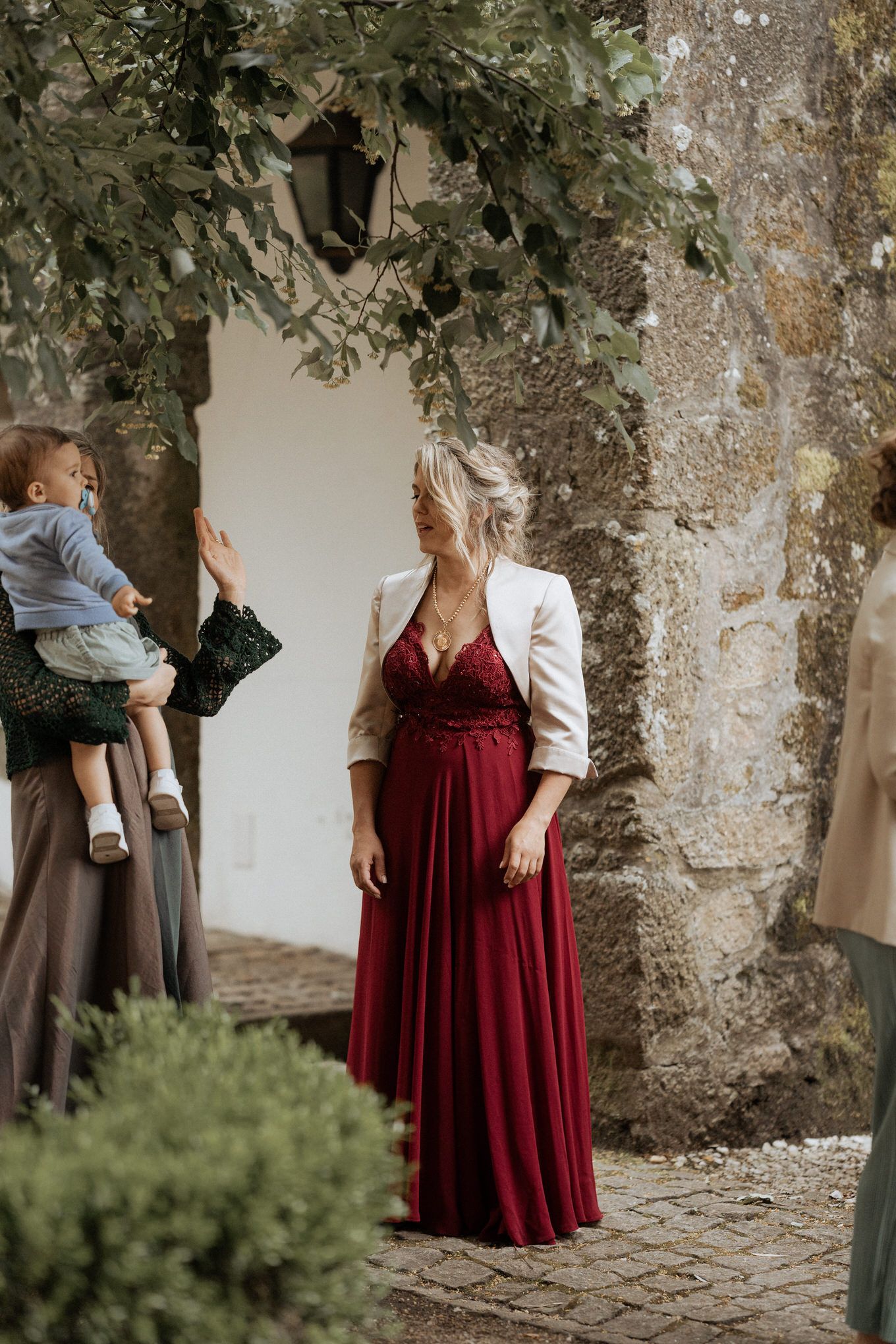 Wedding guest in burgundy gown and cream jacket stands near stone wall with climbing vines.