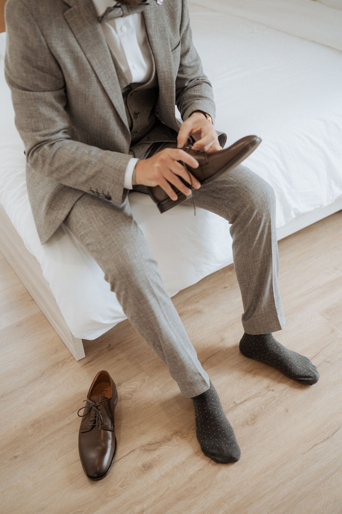 Person in gray suit sitting on white couch putting on dark leather dress shoes with complementing gray socks.