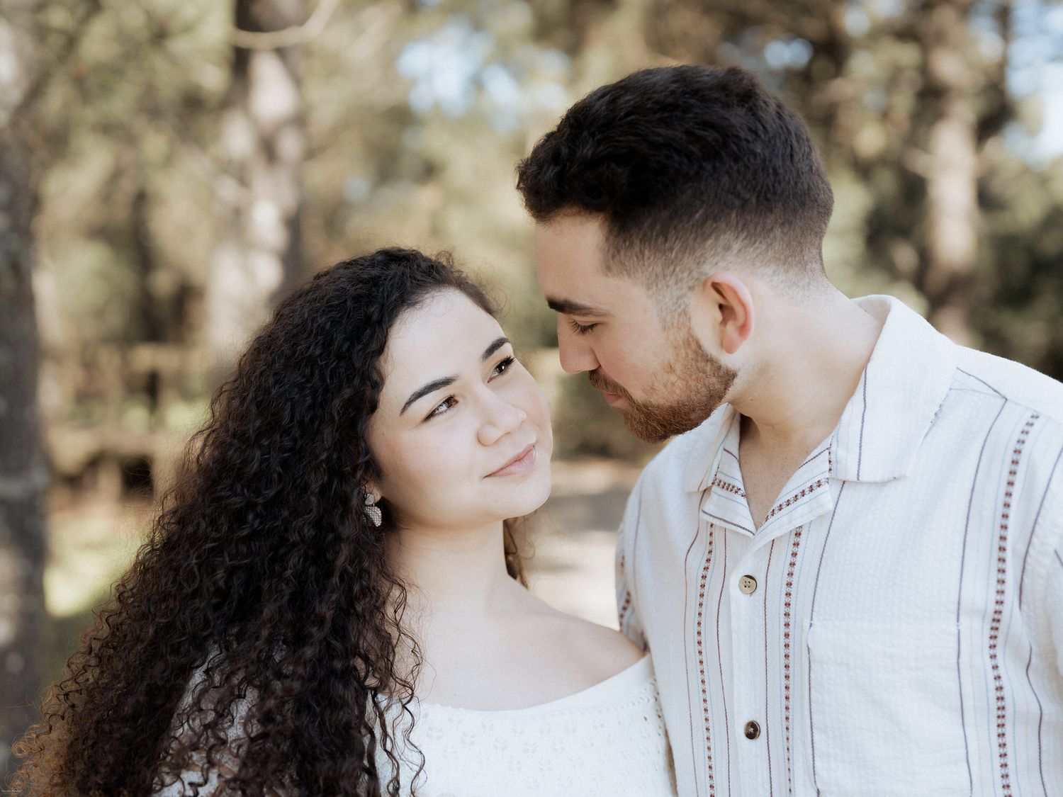 Um casal a olhar-se amorosamente ao ar livre, um deles com cabelo comprido e encaracolado e vestindo roupas brancas.
