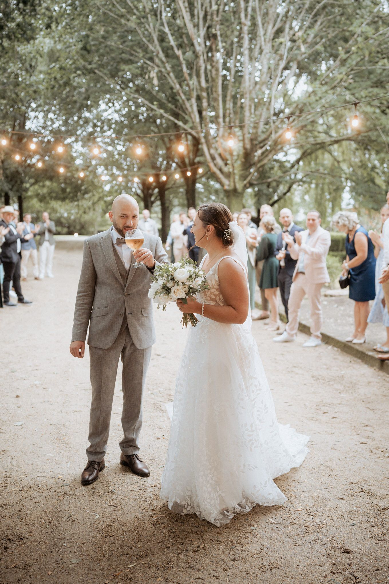 Couple stands together at outdoor wedding reception venue with string lights and trees in background.