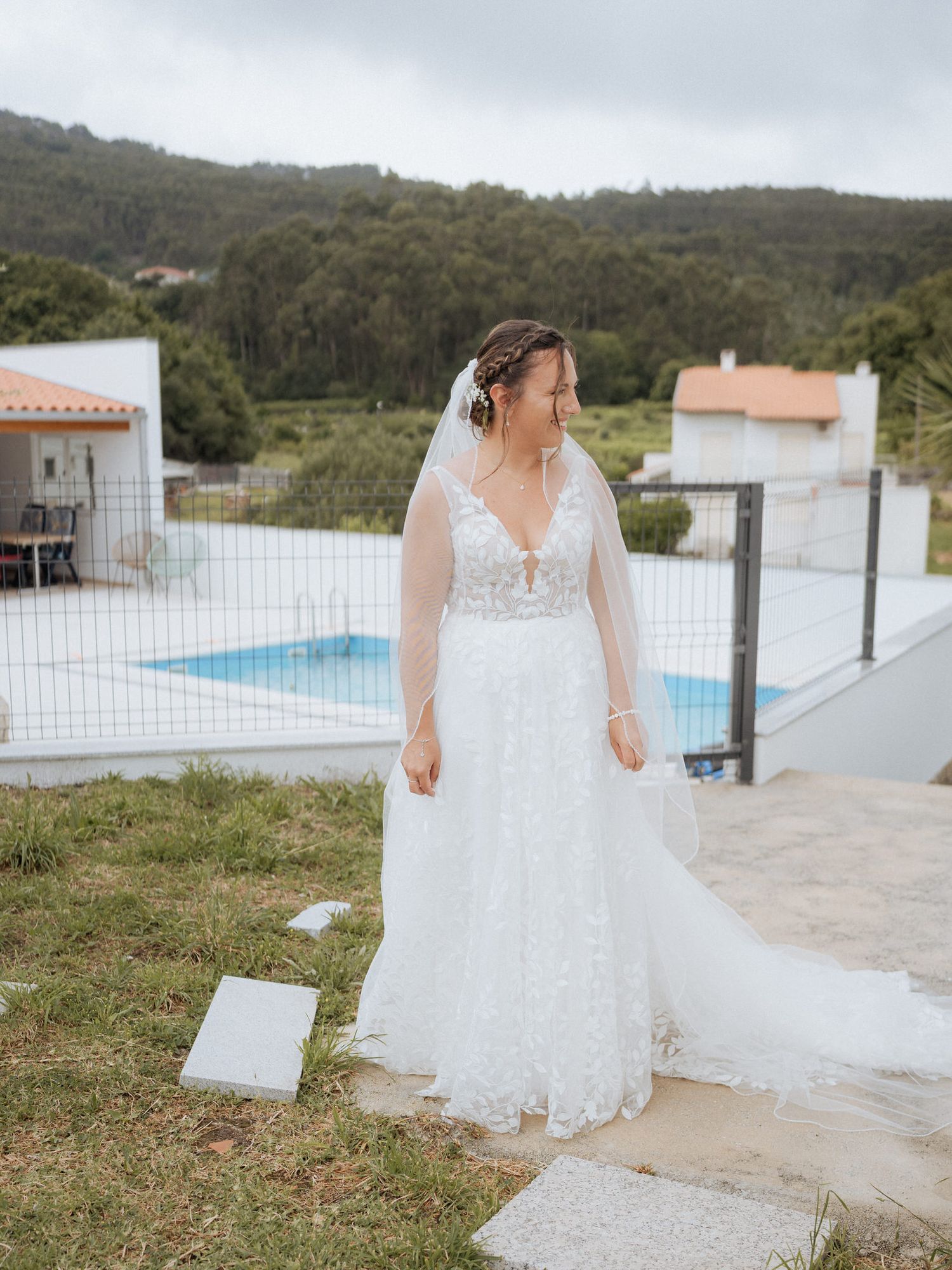 Elegant lace wedding dress with plunging neckline and flowing train shown by pool at outdoor venue with mountain backdrop.