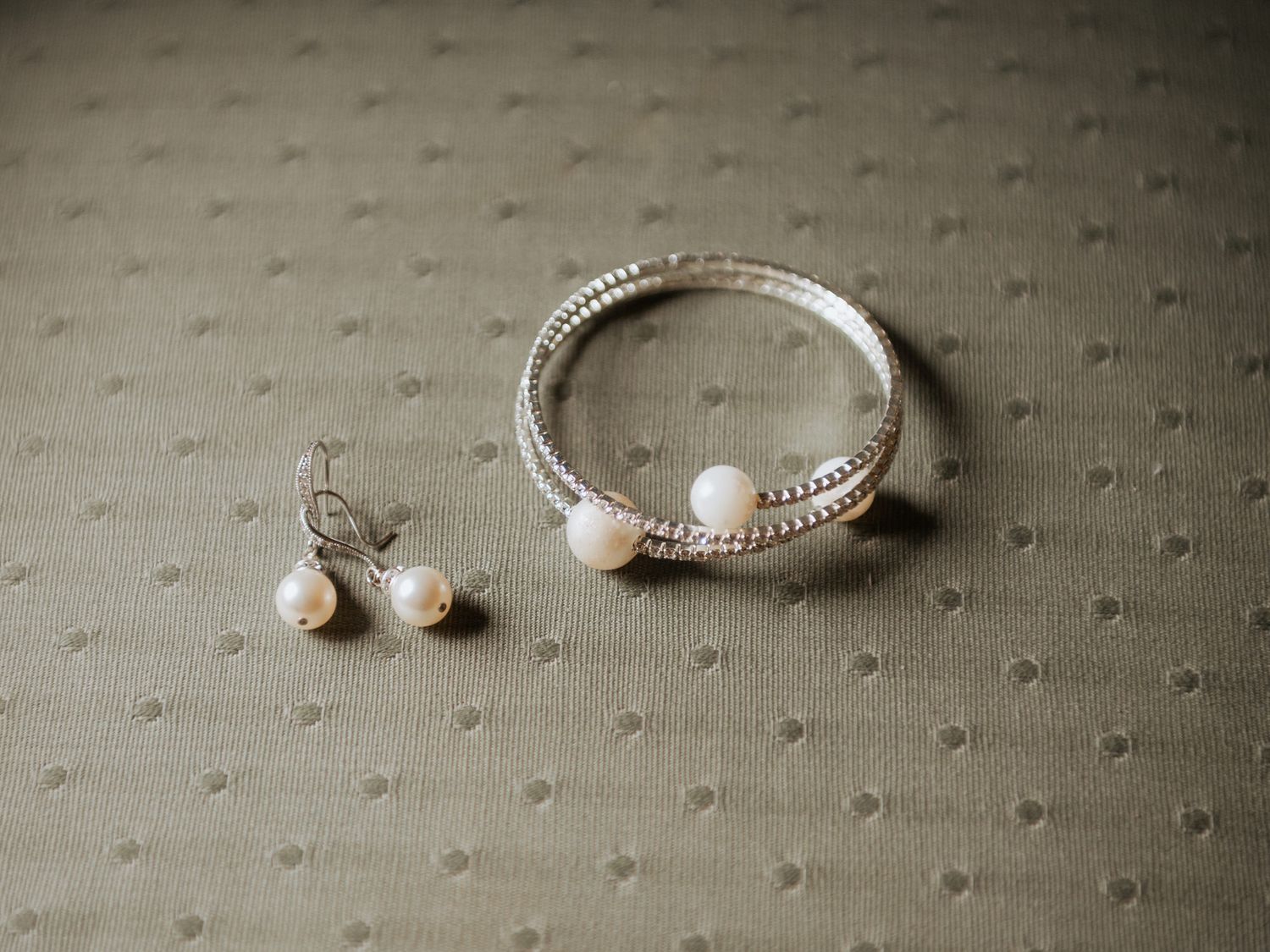 A delicate pearl bracelet and matching stud earrings displayed on a soft sage green textured fabric.