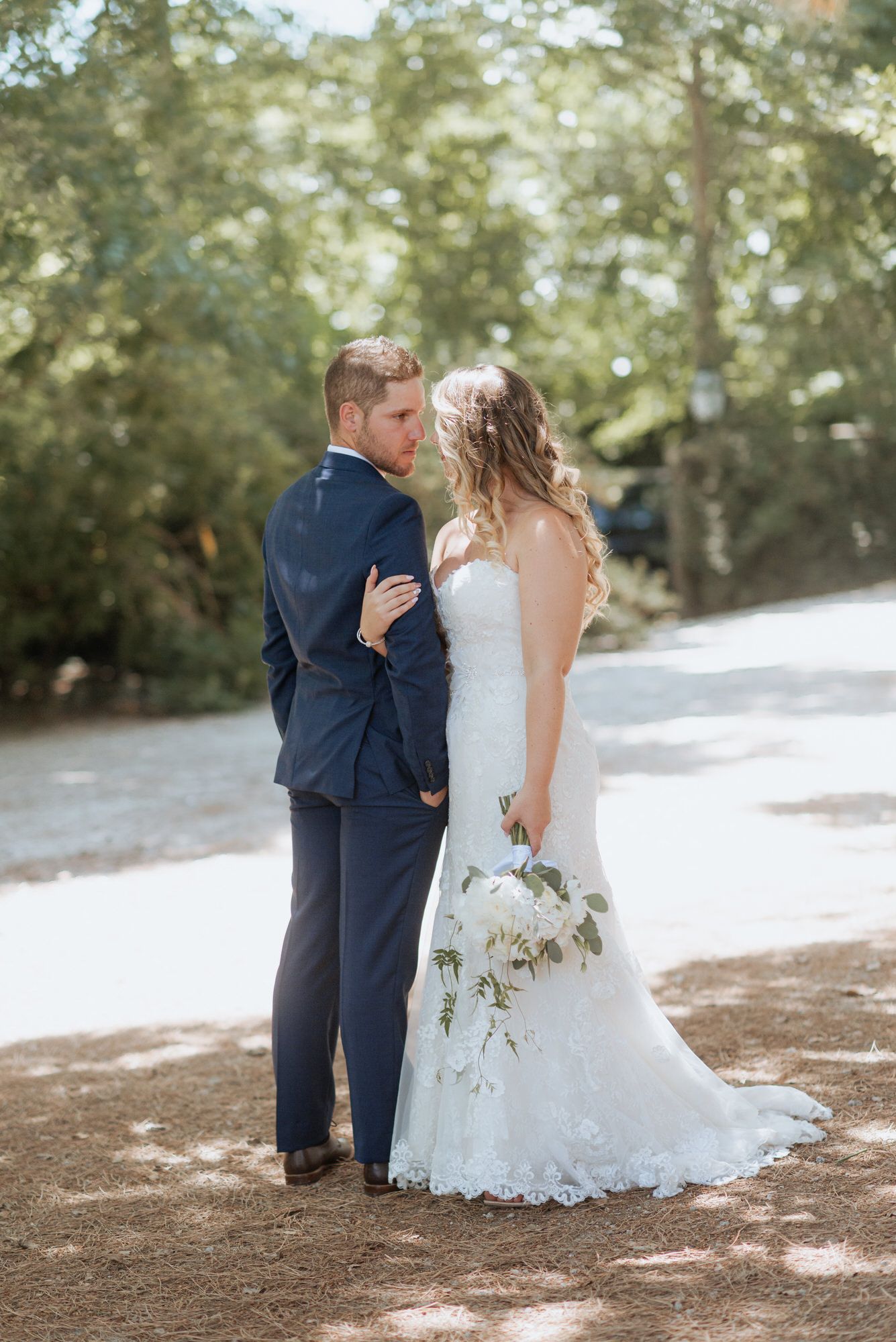 A couple in wedding attire embrace and kiss under a canopy of trees in an outdoor forest setting.