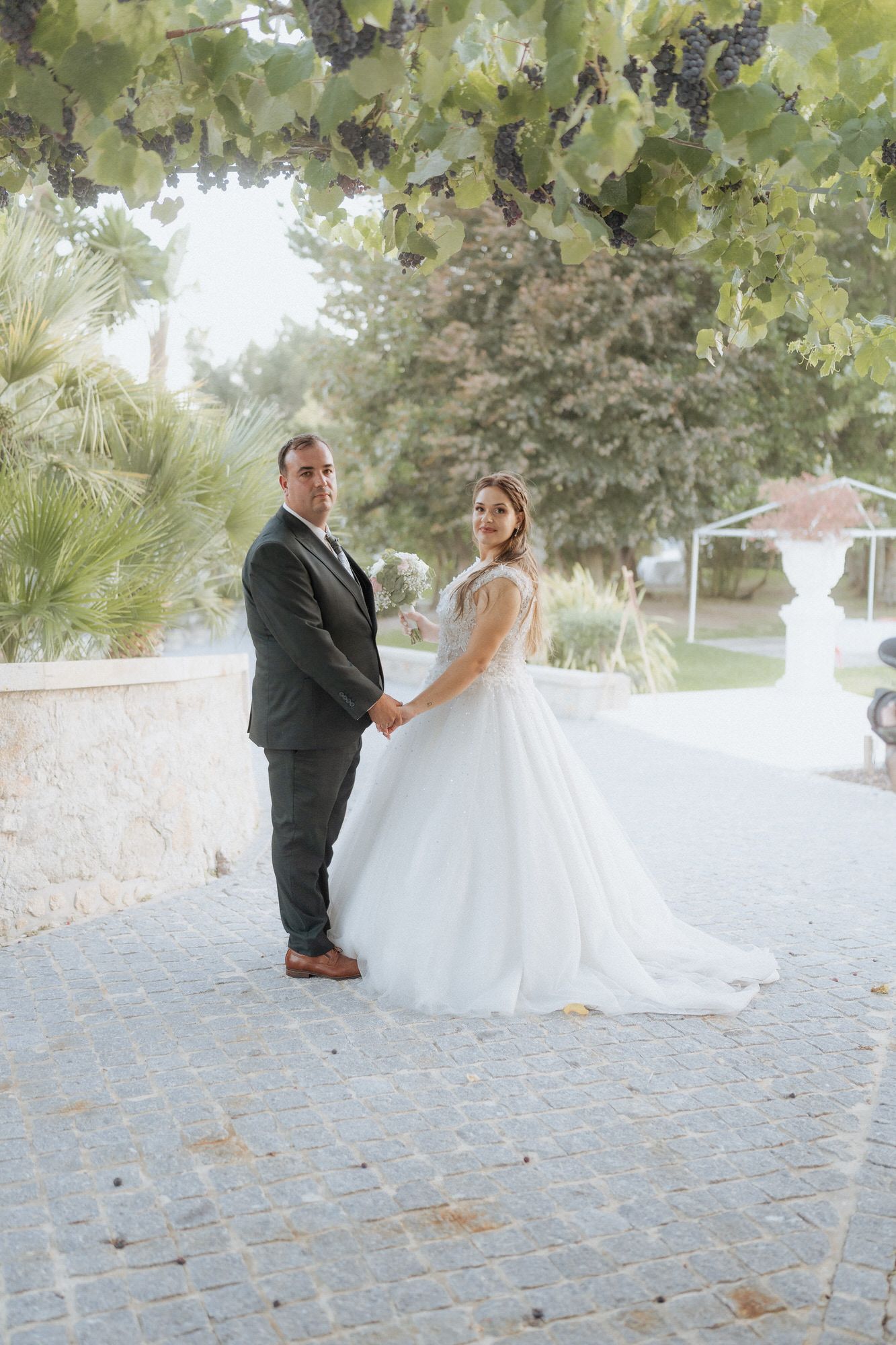 A newlywed couple poses together on a cobblestone path under greenery at their outdoor garden wedding venue.