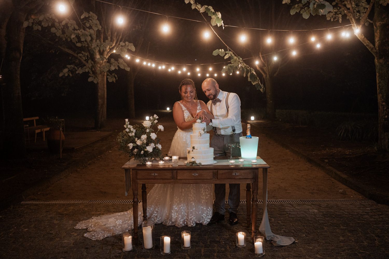 Rustic outdoor wedding cake cutting scene with string lights overhead and floral decorations on wooden table at night.