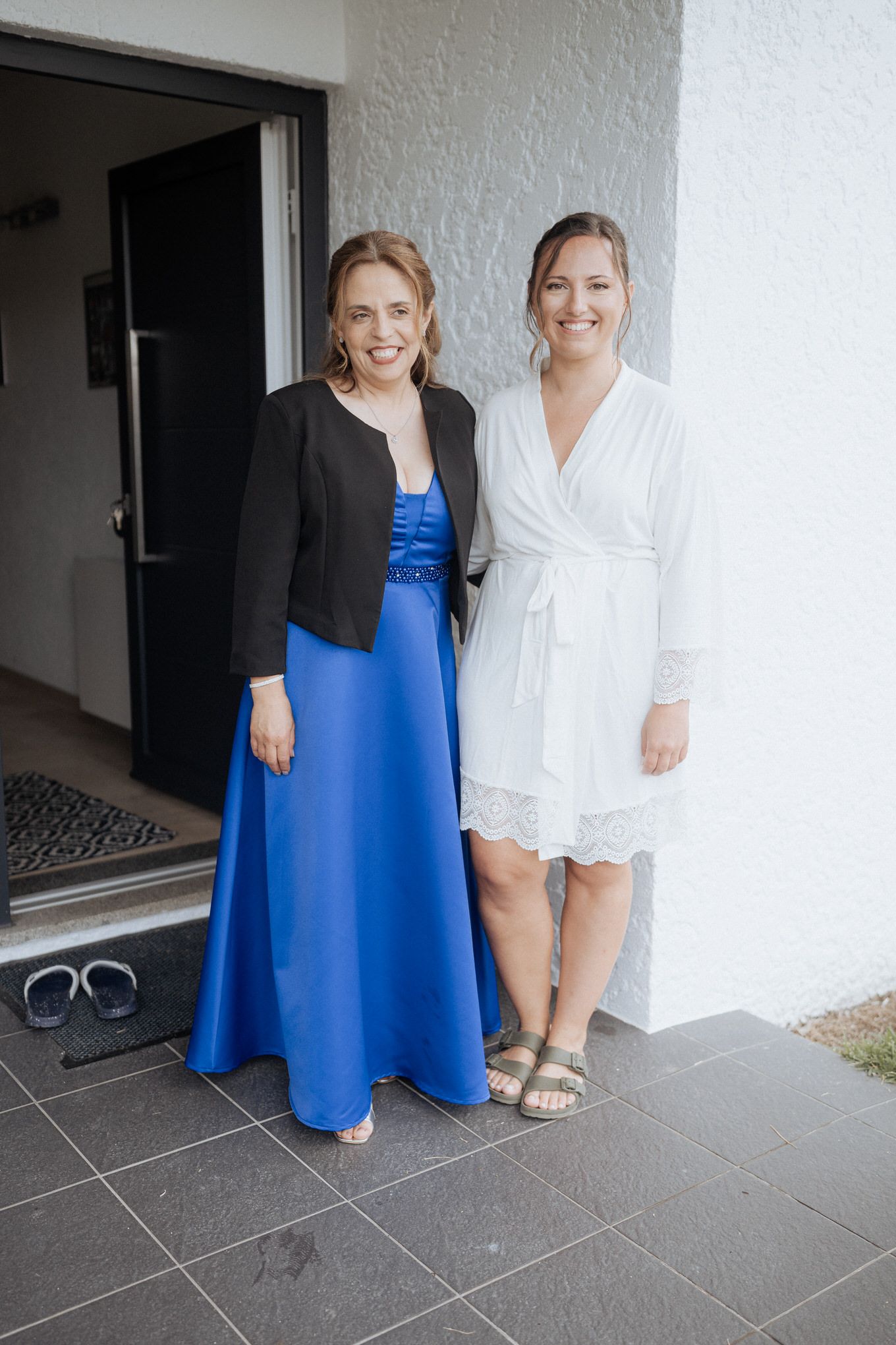 Two women pose together, one wearing a royal blue gown with black jacket and one in a white lace dress.