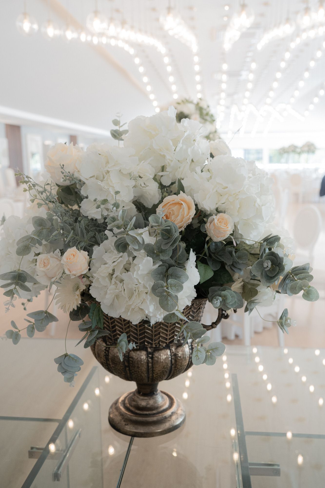Elegant floral centerpiece with white hydrangeas, peach roses and eucalyptus in an antique silver urn on reflective surface.