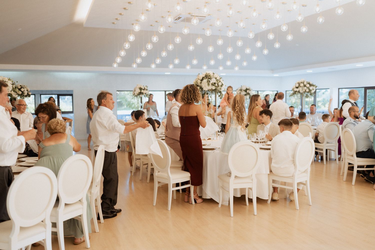 Wedding reception guests stand and celebrate at elegant white venue with modern decor and overhead lighting.