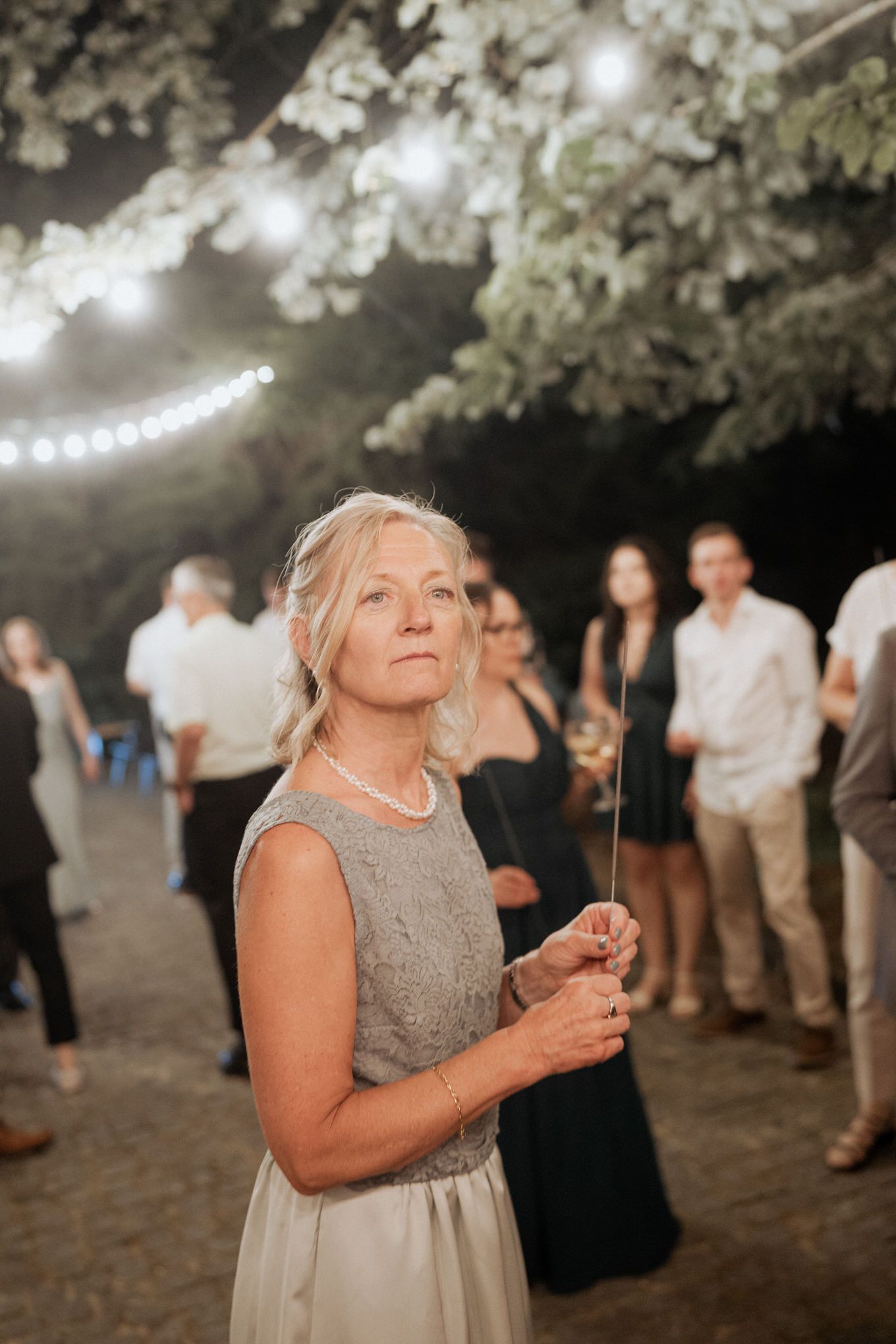 A guest holds a sparkler at an outdoor evening wedding reception under string lights and cherry blossoms.