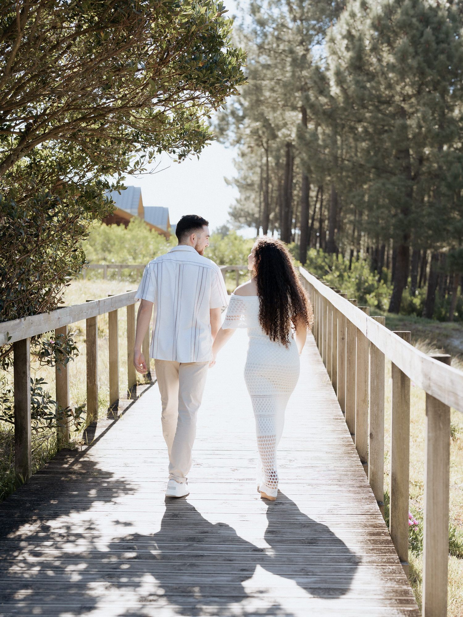 Casal caminhando de mãos dadas em um calçadão de madeira cercado por altos pinheiros em um dia ensolarado.