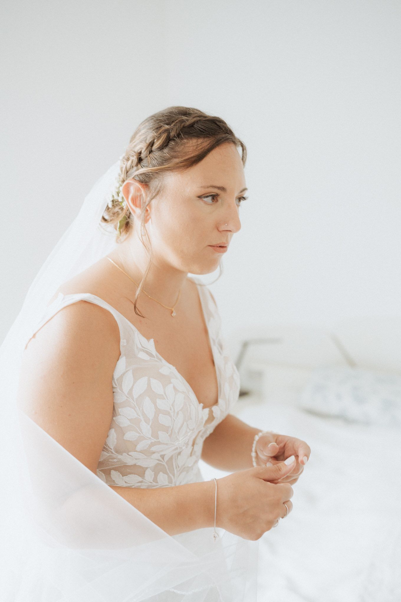 A bride in a white lace wedding dress with braided hair and veil sits on a bright lovely morning.