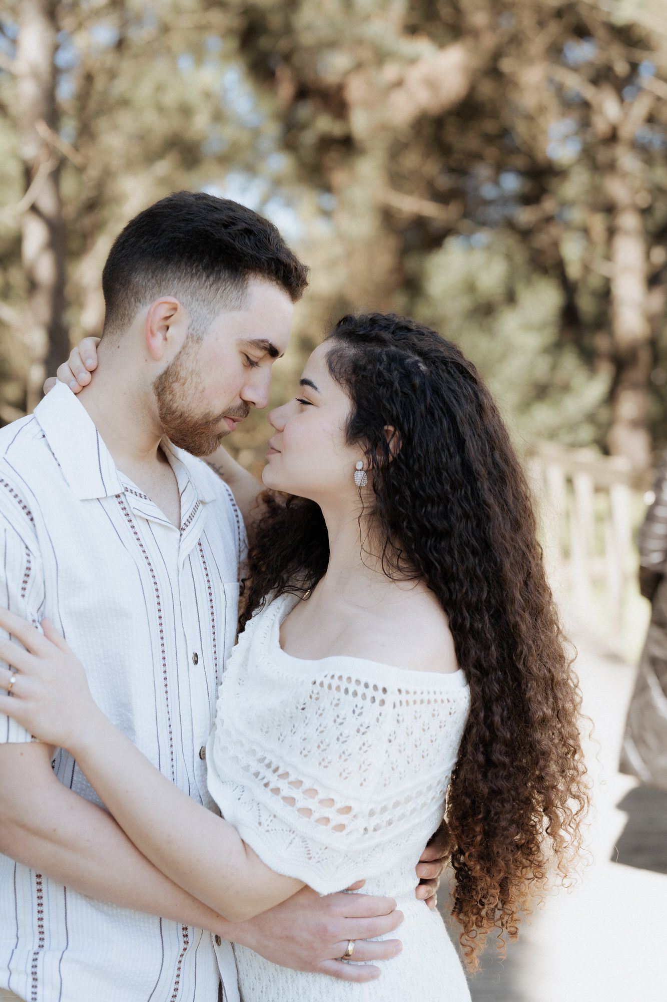 Casal romântico em trajes brancos abraçando-se ao ar livre com árvores ao fundo.