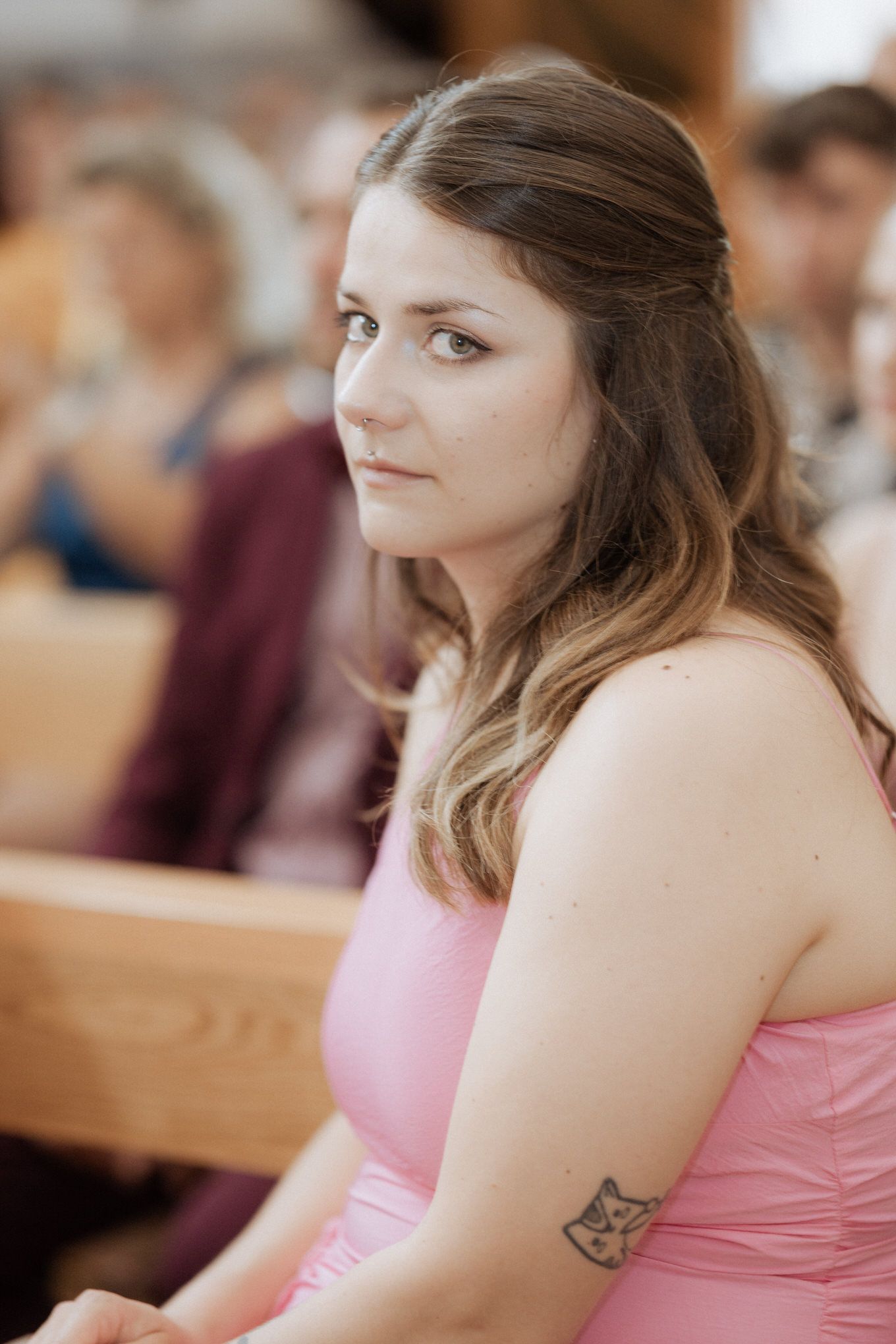 A person in a pink sleeveless dress sits attentively with wavy brown hair and a small tattoo visible on their arm.