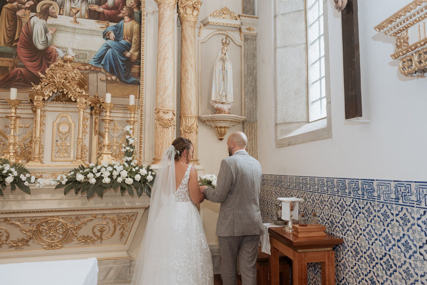 A couple prays at an ornate church altar decorated with white floral arrangements and traditional religious artwork.