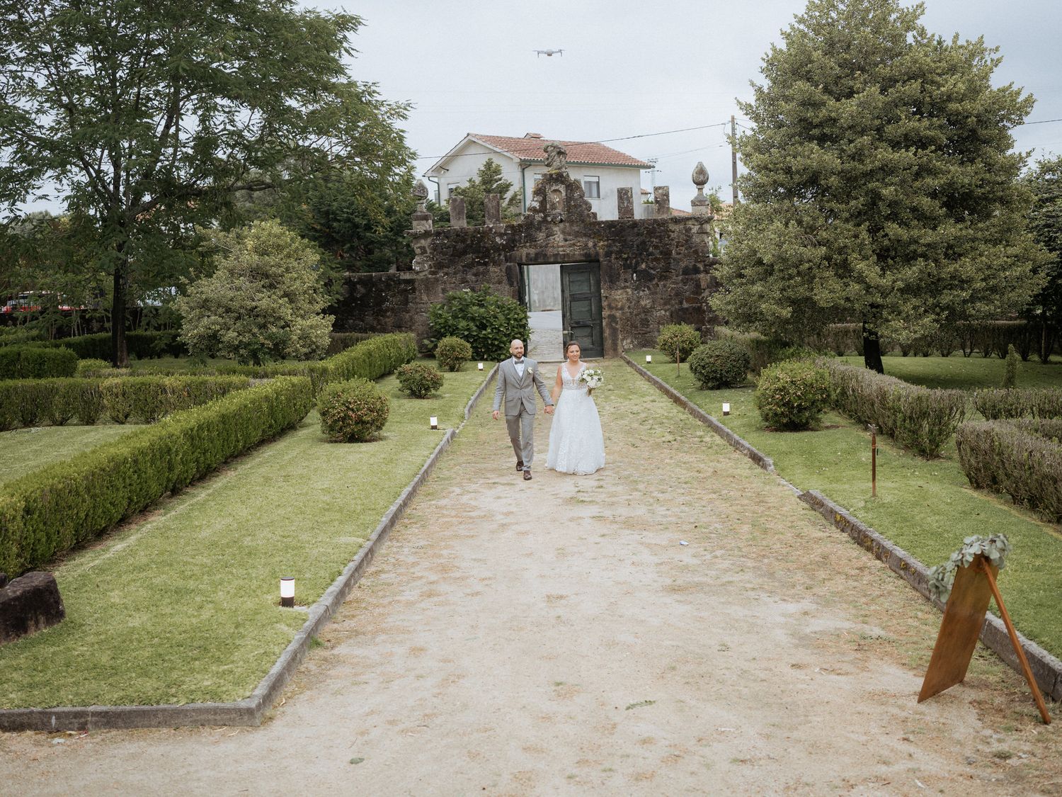 A couple walks down a manicured garden path leading to an elegant white manor house on a sunny day.