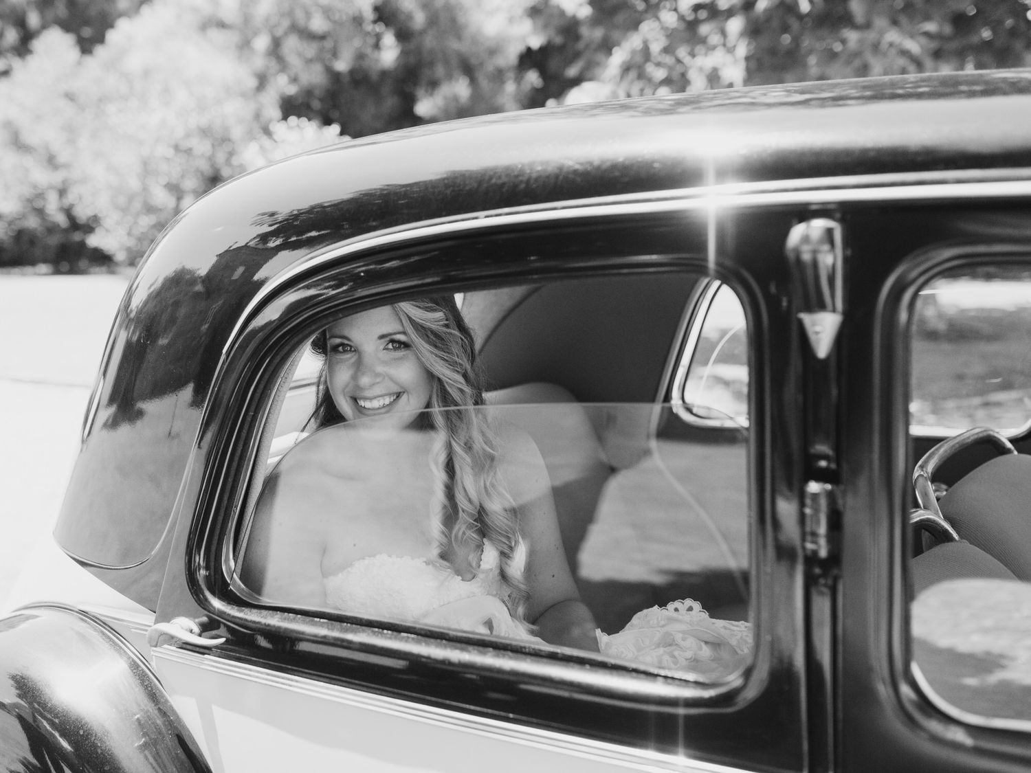 A vintage black and white photo of someone seated in the back of a classic car, viewed through the window.