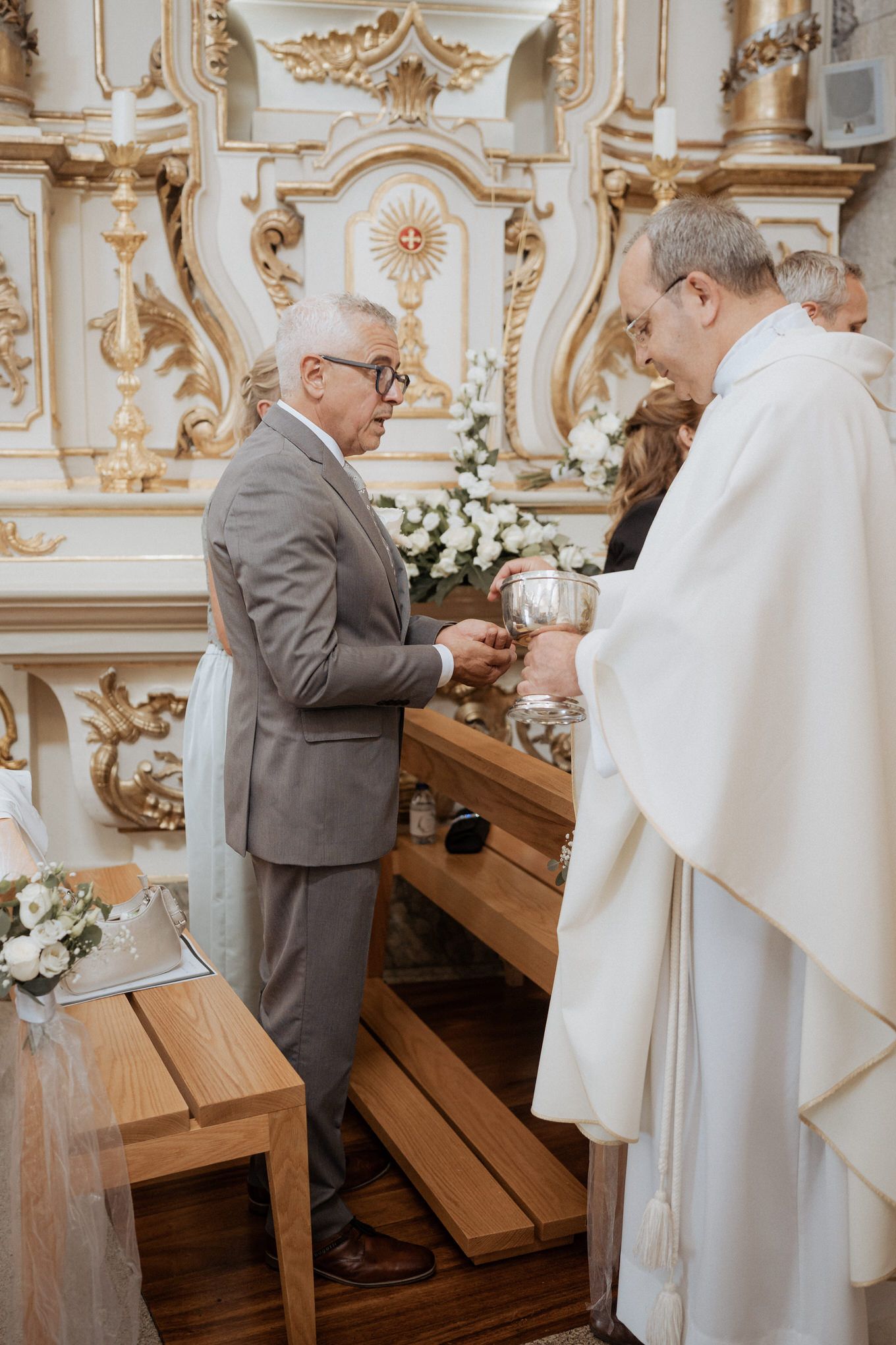 A reverent moment during a Catholic church ceremony with decorative baroque altar and white floral arrangements in the background.
