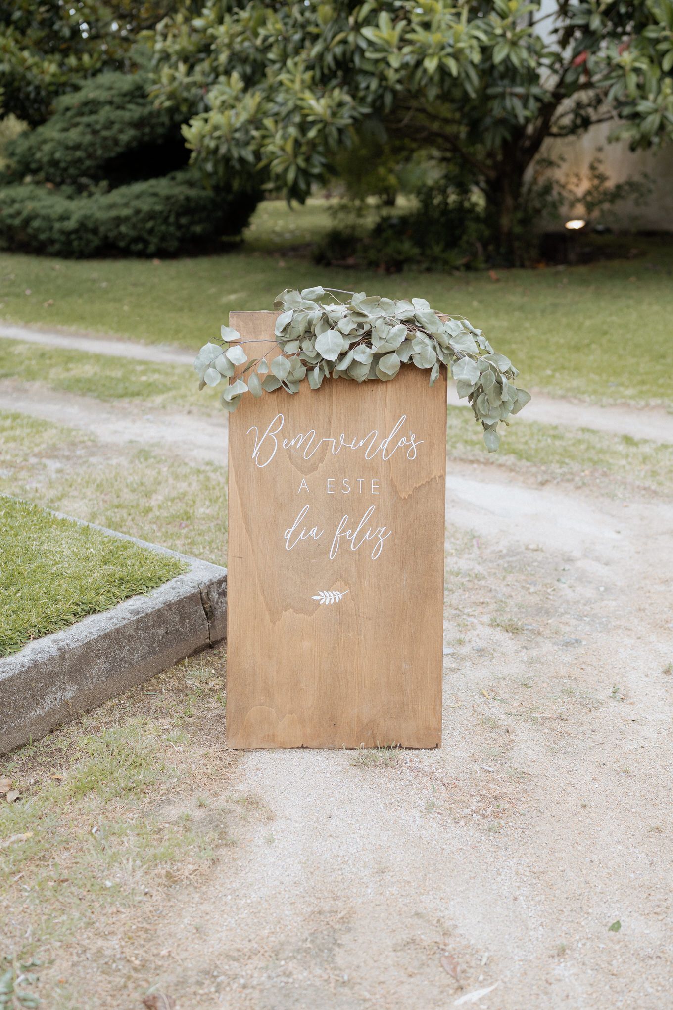 A rustic wooden wedding sign decorated with delicate white flowers and elegant calligraphy text stands on a garden path.