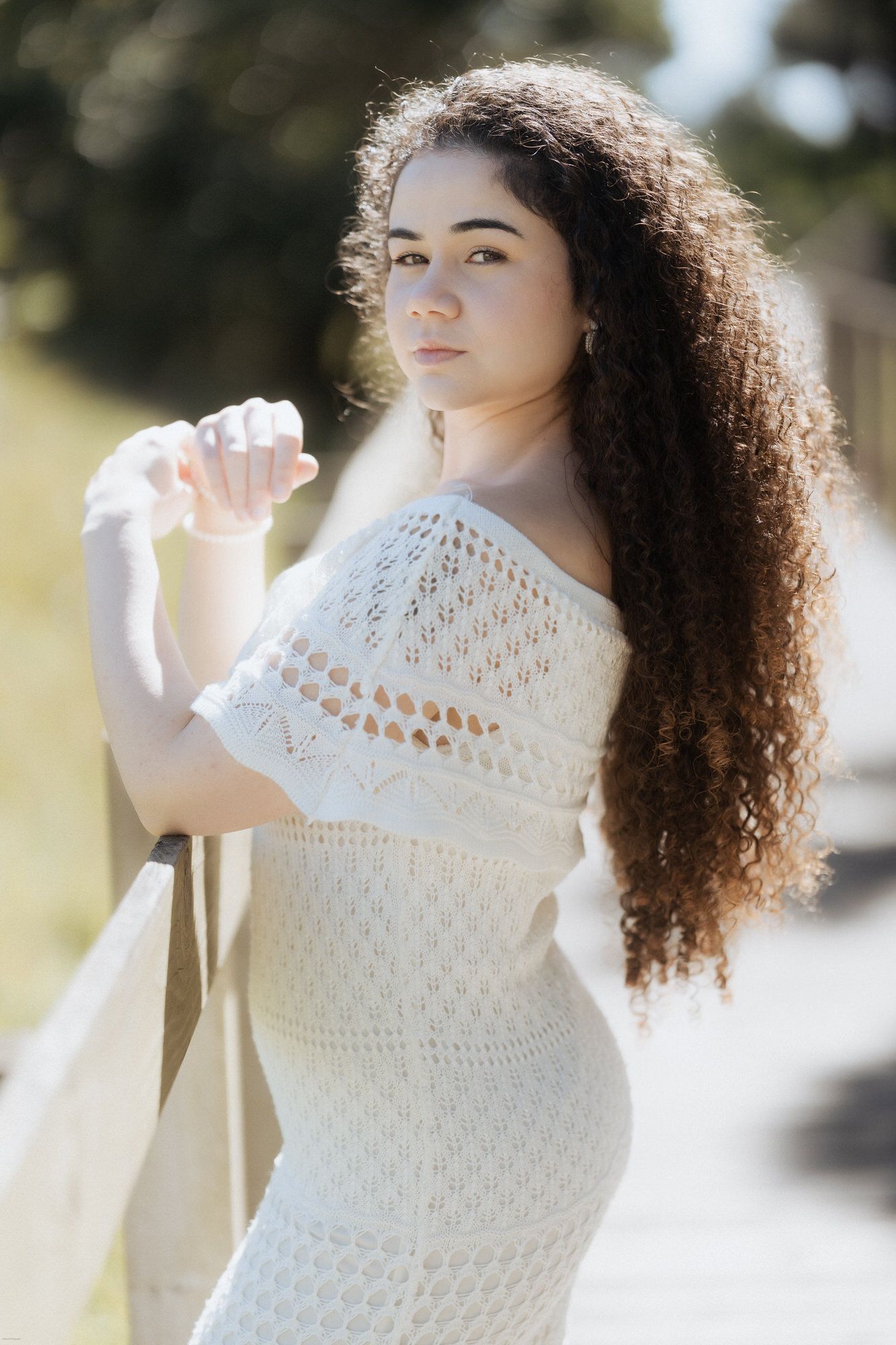 Uma pessoa com um vestido branco de crochê posa ao ar livre sob a luz suave do sol, com longos cabelos cacheados esvoaçando na brisa.