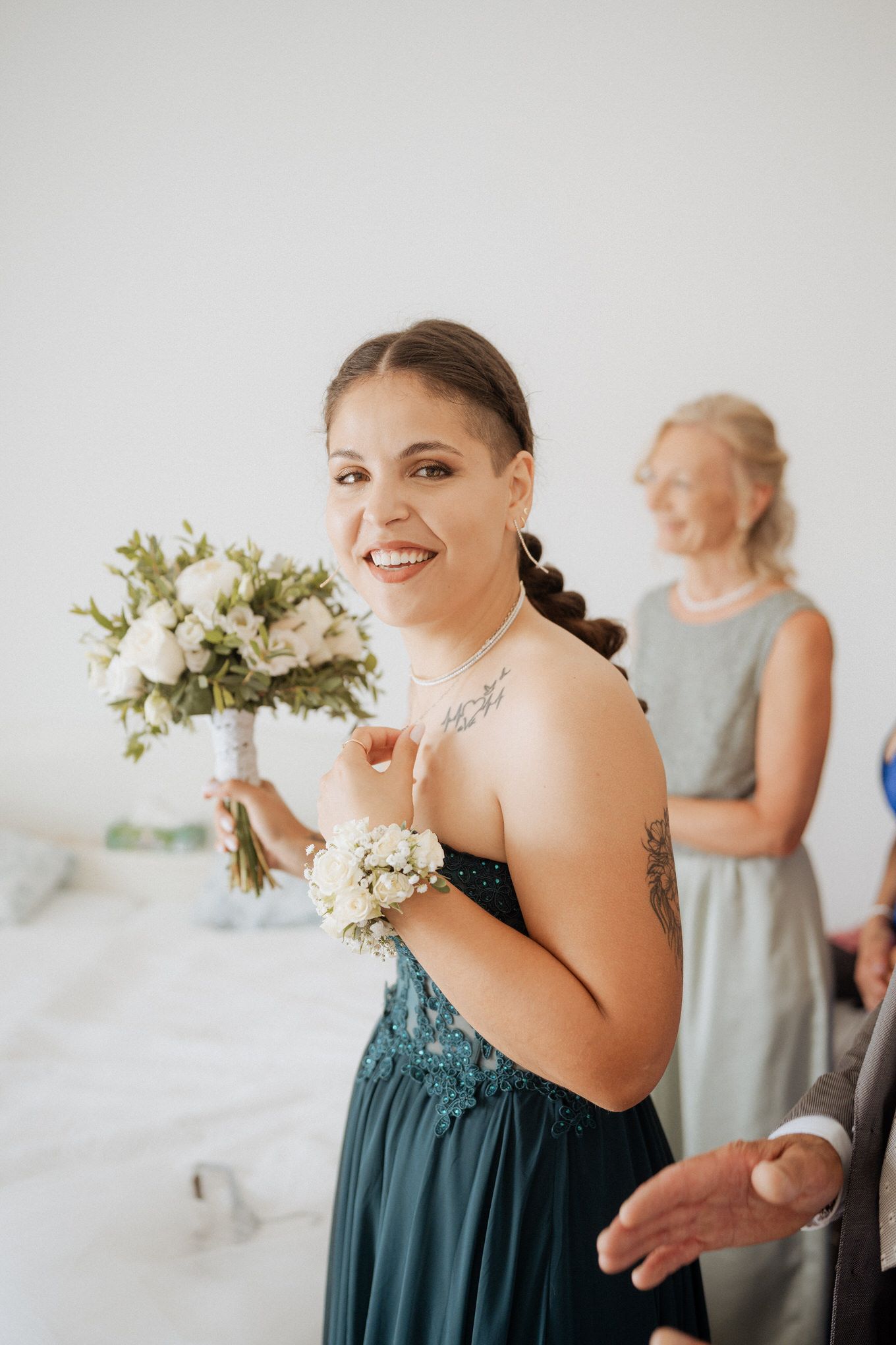 A bridesmaid in a dark teal dress smiles while holding a white floral corsage during wedding preparations.