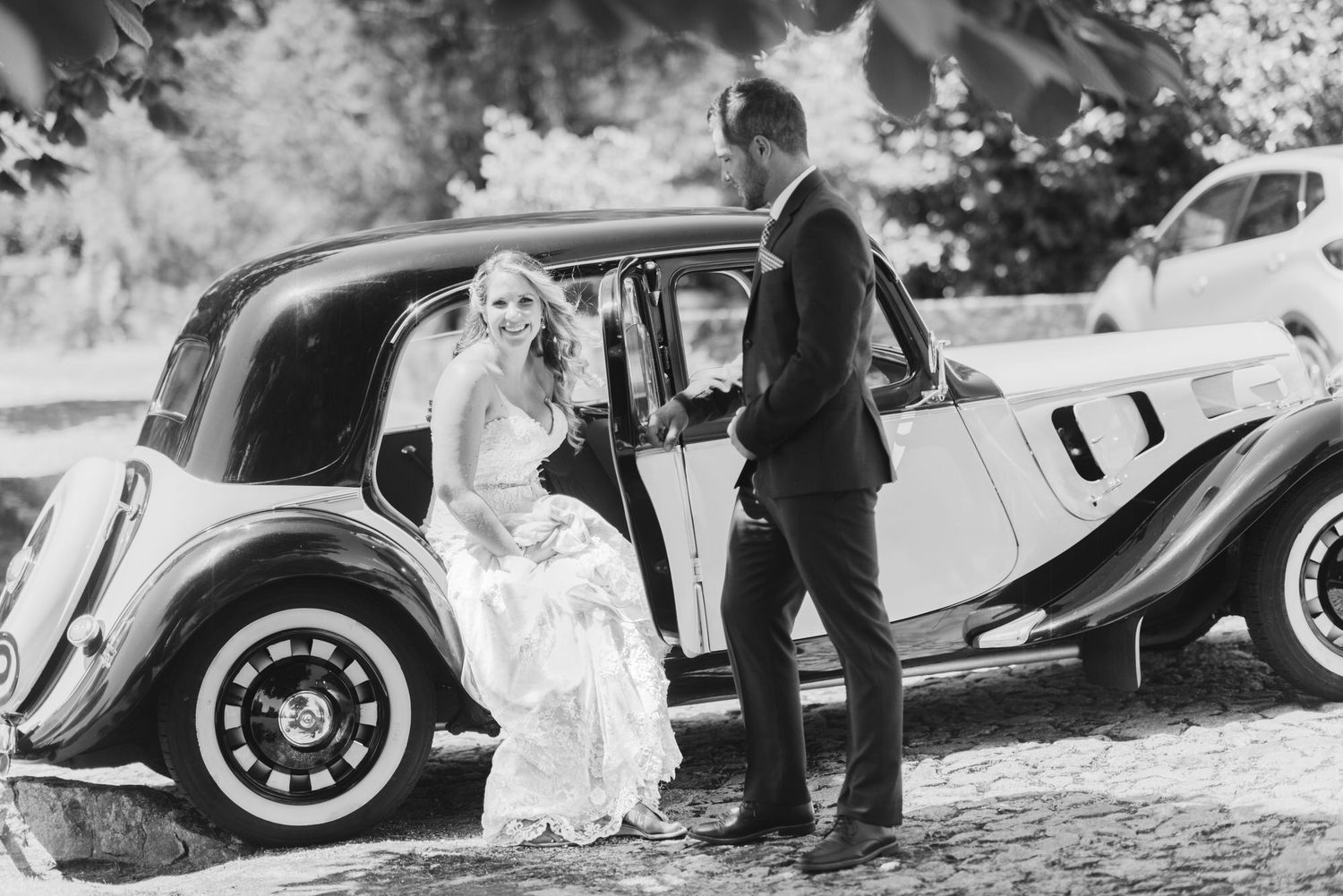 A vintage car serves as an elegant backdrop for a black and white wedding scene on a sunny day.