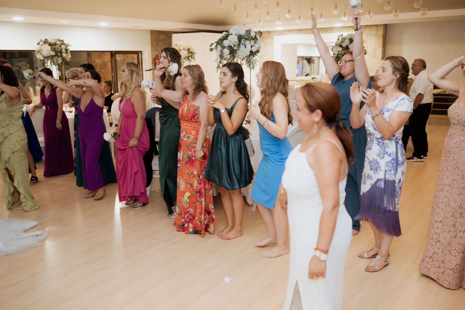 A group of wedding guests in colorful formal attire dance together at a wedding reception on a light-colored dance floor.
